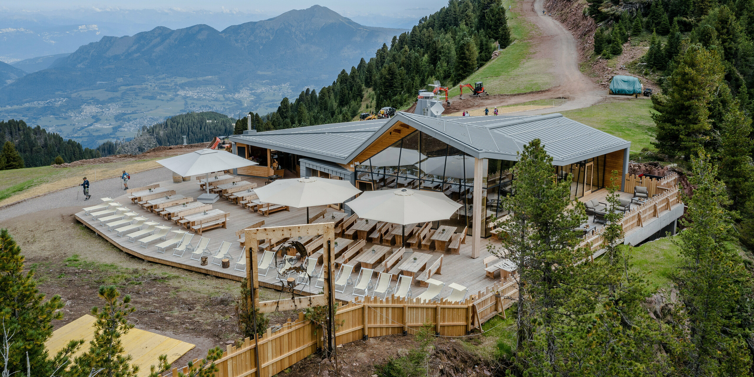 Ansicht des Ristorante Busabelle, erhöht gelegen in einer bergigen Region mit einem spektakulären Blick über das Tal und die umliegenden Berge. Das Gebäude präsentiert eine Kombination aus moderner, hellgrauer PREFALZ Dach- und Fassadenverkleidung sowie großzügigen Holzelementen. Vor dem Restaurant erstreckt sich eine ausgedehnte Terrasse mit Holzmobiliar und Sonnenschirmen, die eine einladende Atmosphäre für Besucher schafft, die auf den Rad- und Wanderwegen unterwegs sind.