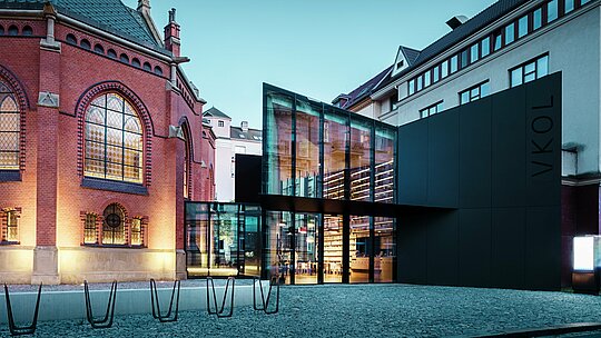 Das Bild zeigt eine beeindruckende Architektur in Olmütz, die die historische Rote Kirche mit einem modernen Neubau verbindet. Die prägnante schwarze Fassade aus PREFABOND Aluminium-Verbundplatten des Neubaus kontrastiert perfekt mit den roten Backsteinmauern der Kirche. Große Glasflächen ermöglichen einen Einblick in das Innere des Neubaus, wo Regale mit Büchern und eine einladende Atmosphäre zu sehen sind. Die präzise Ausführung der Materialien schafft eine harmonische Balance zwischen den historischen und modernen Elementen. Die Verwendung von langlebigen und ästhetisch ansprechenden Materialien wie PREFABOND sorgt nicht nur für eine moderne Optik, sondern auch für dauerhafte Qualität und Funktionalität. Diese Architektur zeigt auf eindrucksvolle Weise, wie Alt und Neu miteinander verschmelzen können, um ein einzigartiges urbanes Ensemble zu schaffen.