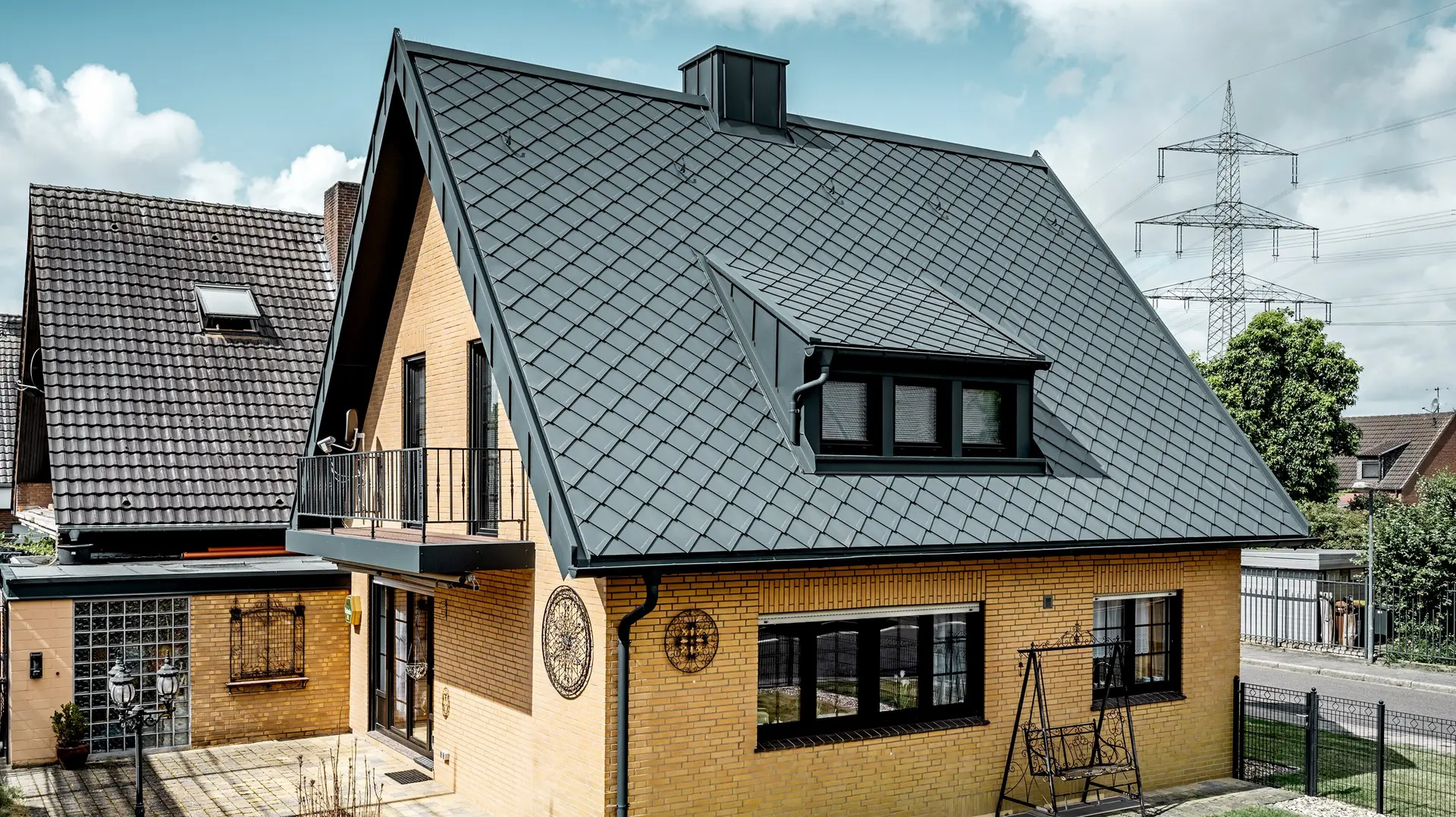Enkeltstående hus i gul klinker etter takrenovering med PREFA takrombe 29 × 29 i mørk grå; nybelagt, bratt sadeltak med rombemønster, integrert takvindu og skorstein, terrasse og hage i forgrunnen.