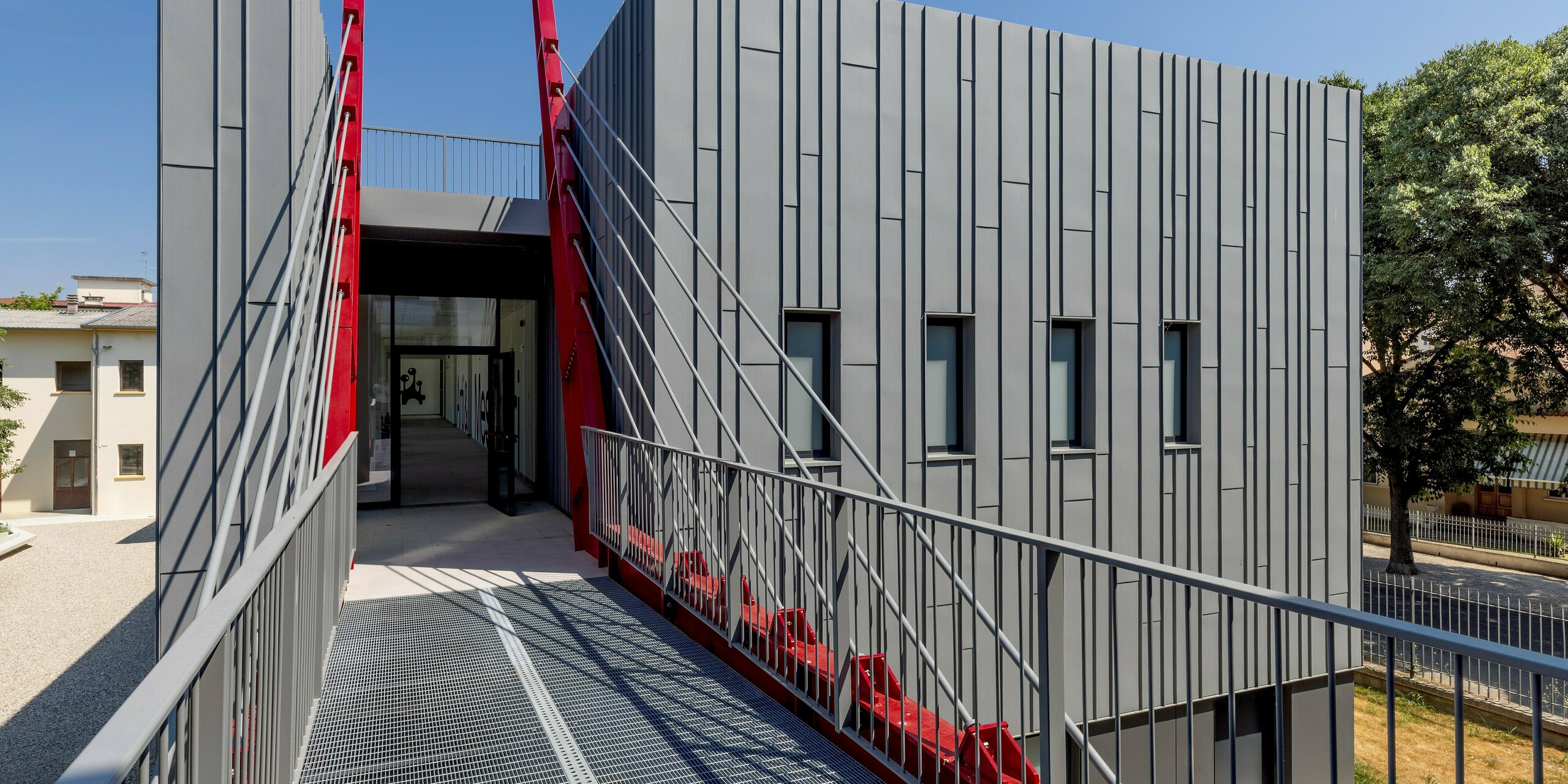 Ein dramatischer Blick auf die Scuola Arte e Mestieri F. Bertazzoni in Suzzara: Eine kühne Brücke mit Stahlgitterrost führt direkt zum Eingang, flankiert von leuchtend roten Diagonalstreben. Diese kontrastieren mit der streng vertikal gegliederten, dunkelgrauen Aluminiumfassade. Die PREFALZ Fassade verleiht dem Bauwerk eine präzise, langlebige Oberfläche mit höchster Witterungsbeständigkeit. Die Bildungseinrichtung beweist, dass sich mit hochwertigen PREFA Aluminiumprodukten spektakuläre Ergebnisse im modernen Design erzielen lassen.