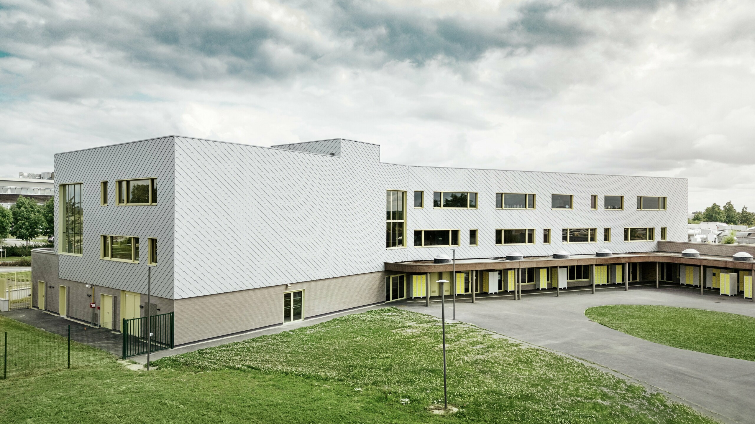 Das Bild zeigt die Rückseite des Collège Léontine Dolivet. Im Vordergrund befindet sich ein großzügiger, gepflasterter Schulhof mit klaren Linien und offenen Flächen, der sich ideal für Pausen und Freizeitaktivitäten eignet. Mehrere Grünflächen lockern die eintönige Umgebung auf. Im Hintergrund erhebt sich ein modernes Schulgebäude mit einer markanten Aluminiumfassade aus silbermetallischen PREFA-Wandelementen, die das Licht reflektieren und so eine dynamische Wirkung erzeugen. Zahlreiche große Fenster lassen viel Licht ins Innere. Die Aufnahme entstand tagsüber bei leicht bewölktem Himmel, wodurch die Metalloberfläche besonders gut zur Geltung kommt.