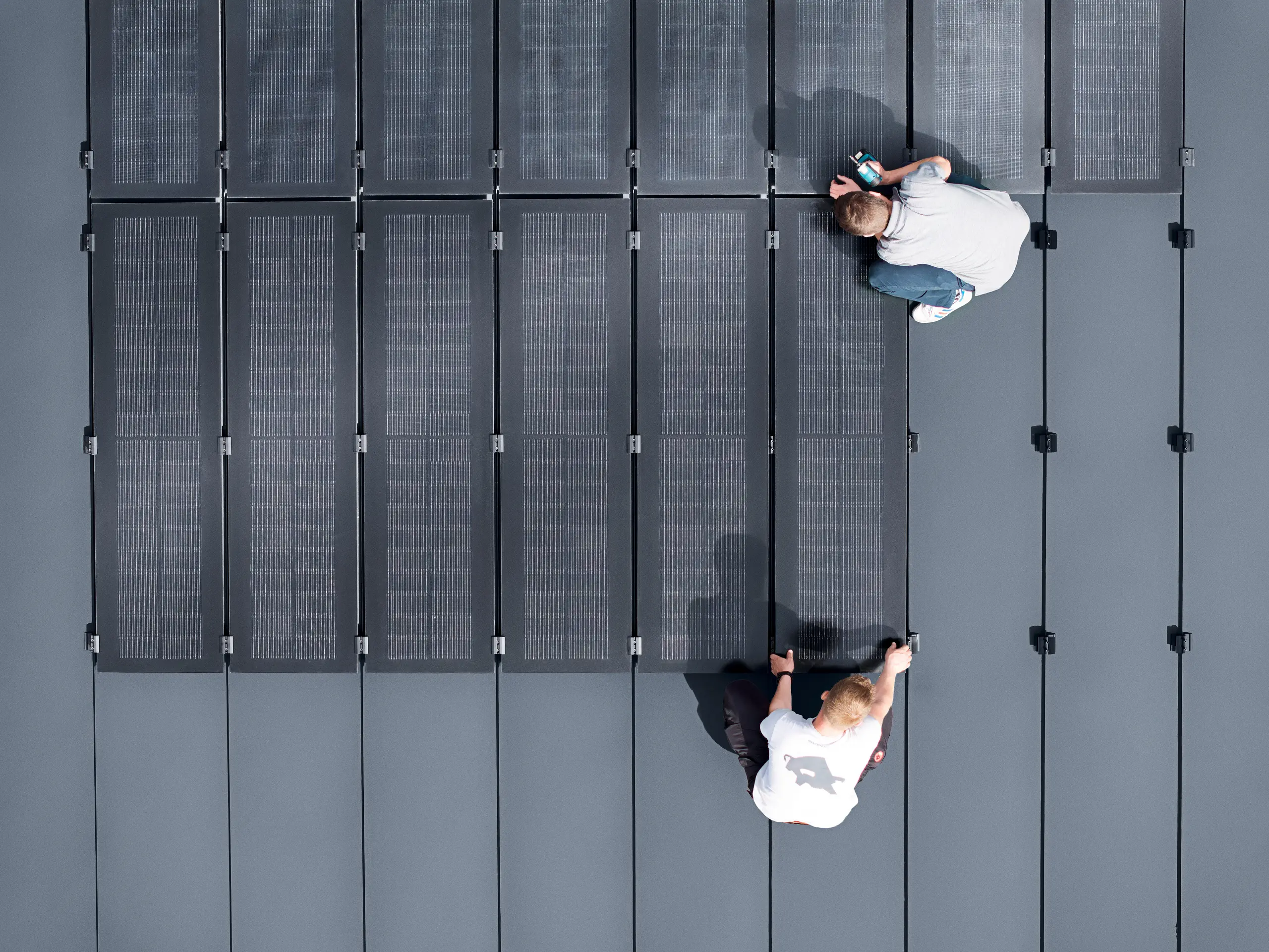 Zwei Personen installieren PREFALZ-Solarmodule auf einer großen grauen Dachfläche, die mit doppelflanschigen Paneelen bedeckt ist.