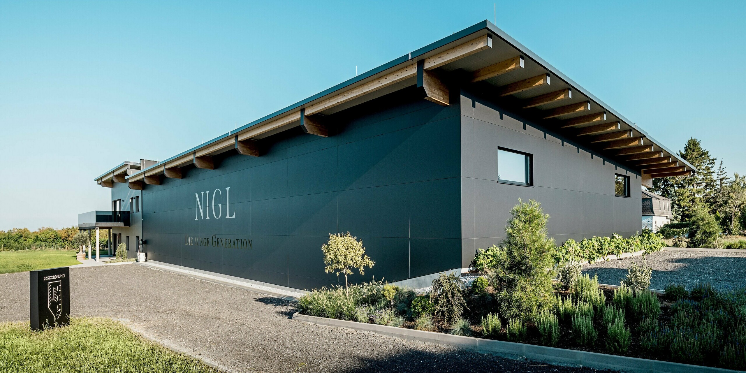 Gesamtansicht der Rückseite des Weinguts Nigl in Perchtoldsdorf mit moderner PREFA Aluminiumfassade in P.10 Schwarz. Die großflächige, glatte Oberfläche der Verbundplatten wird durch klare Fugenlinien gegliedert und trägt prominent den Schriftzug „NIGL – Die junge Generation“. Das weit auskragende Pultdach mit offenen Holzbalken unterstreicht die horizontale Linienführung der Architektur. Seitlich rahmt eine gepflegte, naturnahe Bepflanzung das Gebäude ein. Die PREFA Fassade verbindet moderne Optik mit hoher Witterungsbeständigkeit, Langlebigkeit und nahezu wartungsfreier Funktionalität.