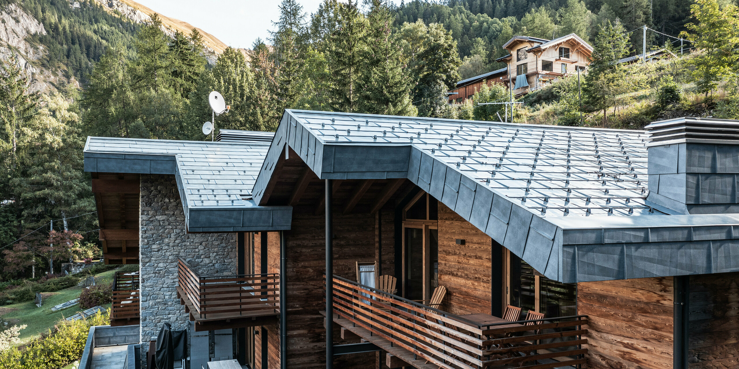 Komplexe Villa in Courmayeur mit Holzfassaden und PREFA Aluminiumpaneele FX.12 in der Farbe P.10 Steingrau. Die Architektur kombiniert moderne und traditionelle Elemente wie Natursteinmauerwerk und mehrstöckige Holzstrukturen, die durch stilvolle Blechdetails ergänzt werden. Mehrere Balkone und Terrassen bieten einen großzügigen Außenbereich, der durch üppiges Grün und die dramatische Berglandschaft ergänzt wird.