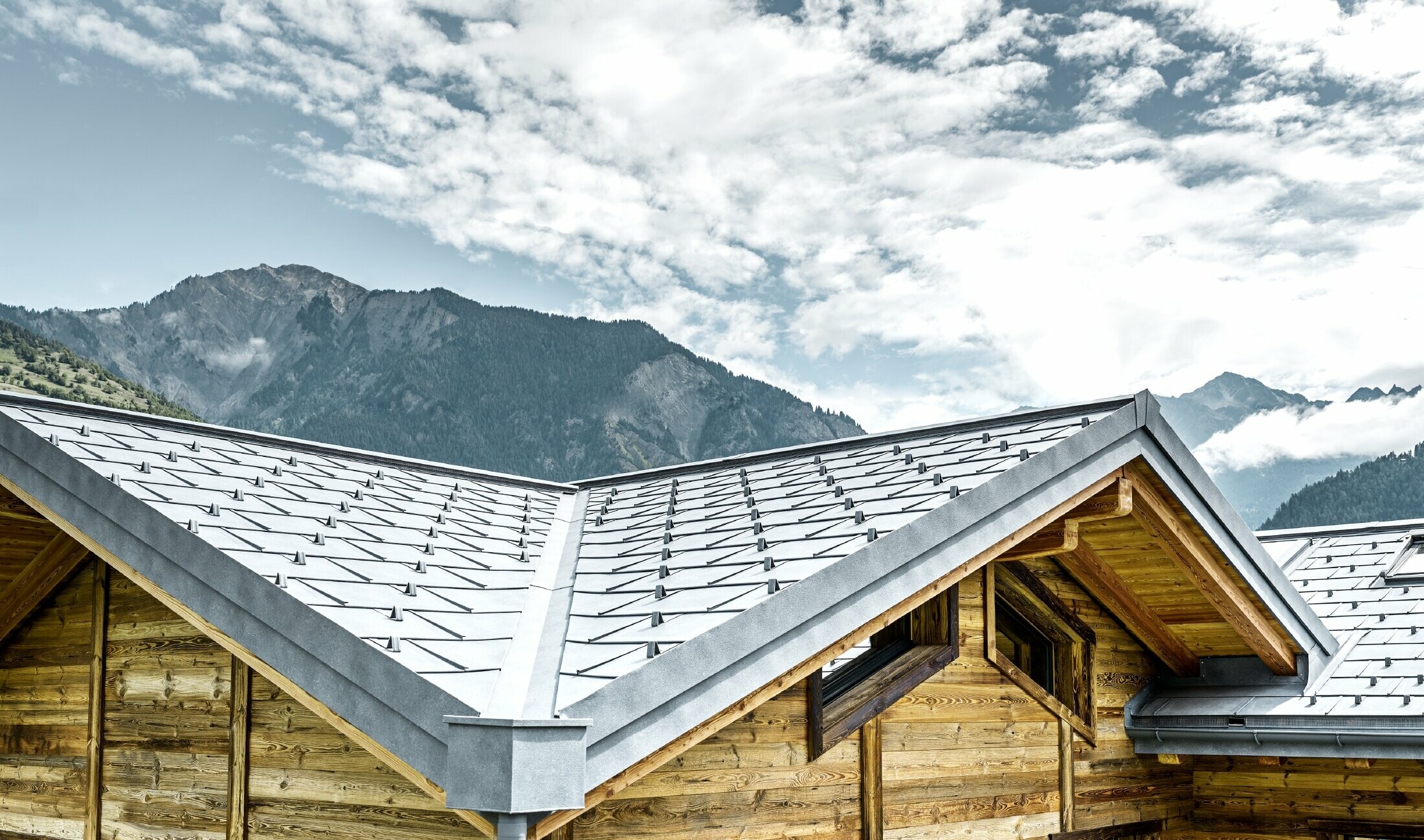 Taket på en rustikk hytte i Sveits med trefasade og aluminiumstak fra PREFA. Takplate R.16 i er lagt. Man ser det vinklede taket og kanalen.