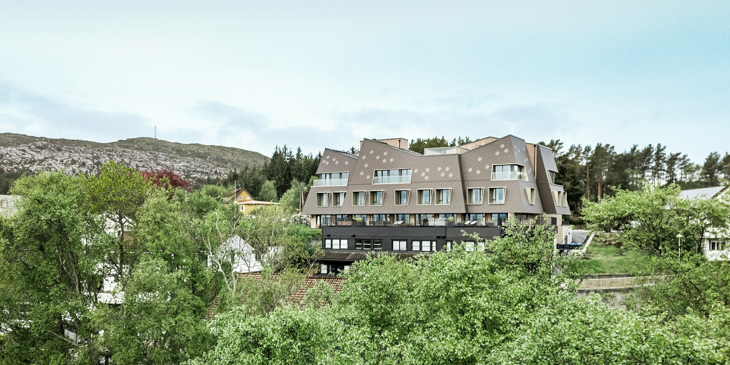 Utsikt på lang avstand fra boutiquehotellet Beckerwyc House i Bekkjarvik i Norge, som ligger i et grønt, kupert landskap. Den futuristisk utseende bygningen er kledd med PREFA-veggfliser 44 × 44 i valnøttbrun, som smelter harmonisk inn i de steinete omgivelsene. Hotellets uvanlige arkitektur skiller seg ut fra de omkringliggende tradisjonelle husene, samtidig som den passer godt inn i naturen takket være den jordnære fargeskalaen.