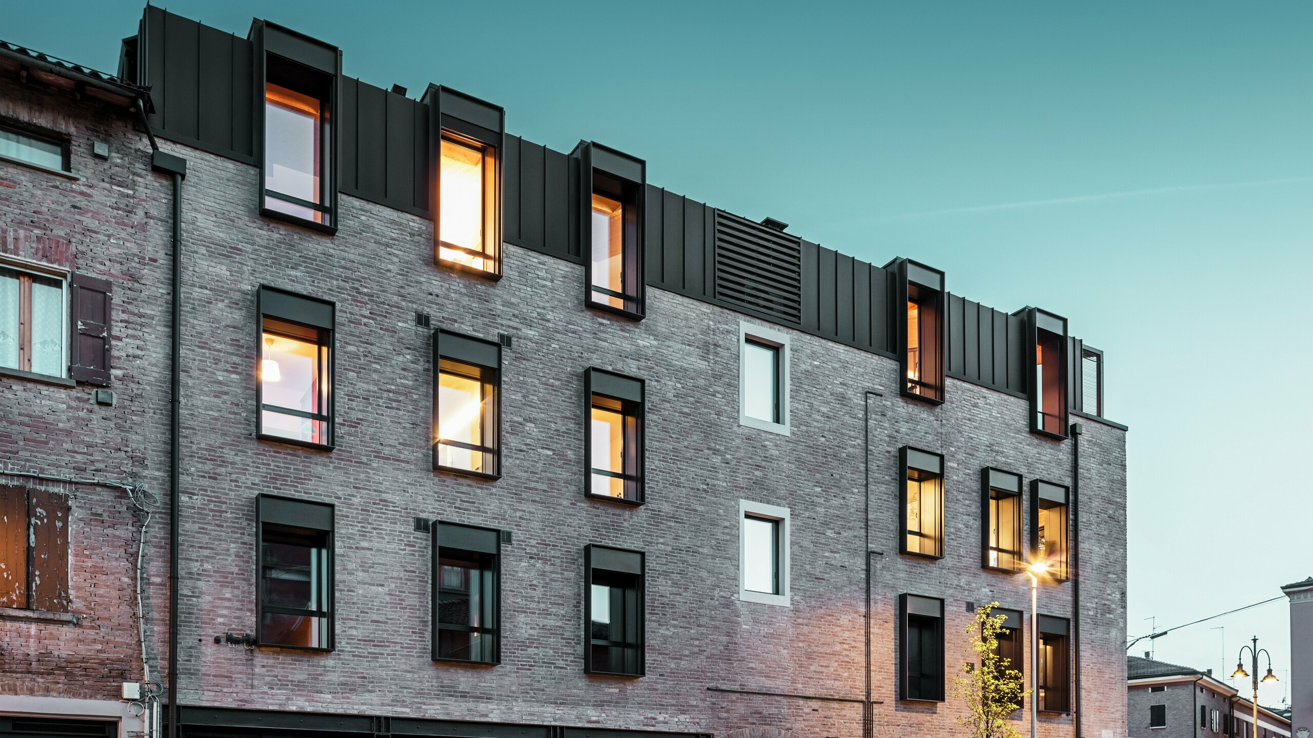 Städtisches Backsteingebäude in Italien mit modernem PREFALZ Aluminiumdach in P.10 Nussbraun, beleuchteten Fenstern und umliegender historischer Architektur in der Abenddämmerung.