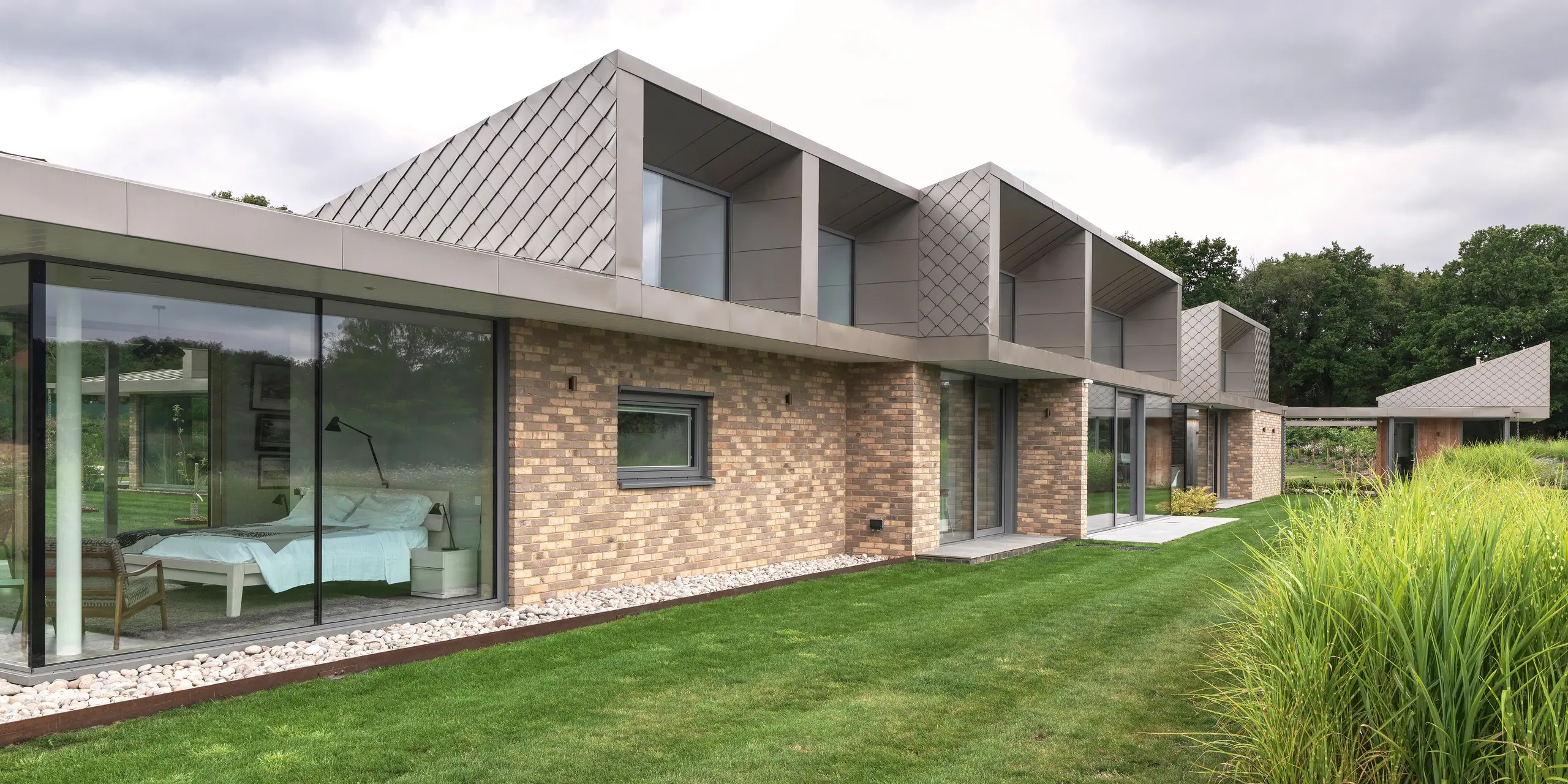 Seitenansicht des modernen Wohnhauses in West Sussex mit markanten Aufbauten im Obergeschoss, verkleidet mit PREFA Wandrauten 20 × 20 in P.10 Bronze. Das flach geneigte PREFALZ Aluminiumdach bildet einen eleganten und witterungsbeständigen Abschluss. Großflächige Verglasungen im Erdgeschoss verbinden Innen- und Außenraum nahtlos und verleihen dem Gebäude ein hochwertiges, zeitloses Erscheinungsbild.