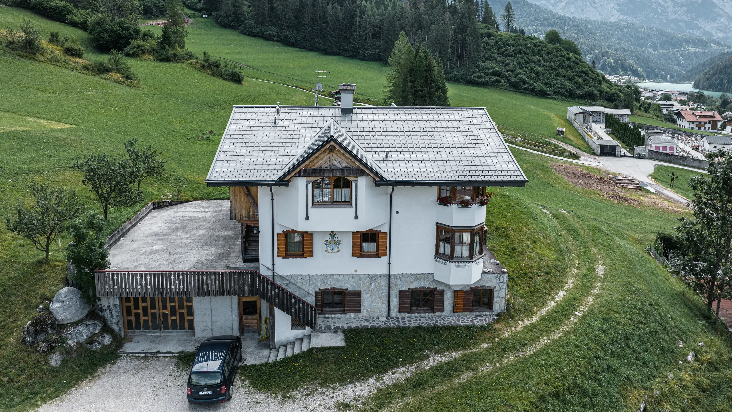 Fugleperspektiv av inngangen til den tradisjonelle fjellhytta i Auronzo di Cadore etter takfornyelsen med PREFA aluminiumsplater i P.10 lys grå. Bygningen med sin karakteristiske alpine sjarm og moderne blikktak føyer seg harmonisk inn i det frodige grønne landskapet og det imponerende fjellpanoramaet i bakgrunnen, og viser en vellykket kombinasjon av funksjonalitet og regional arkitektur.