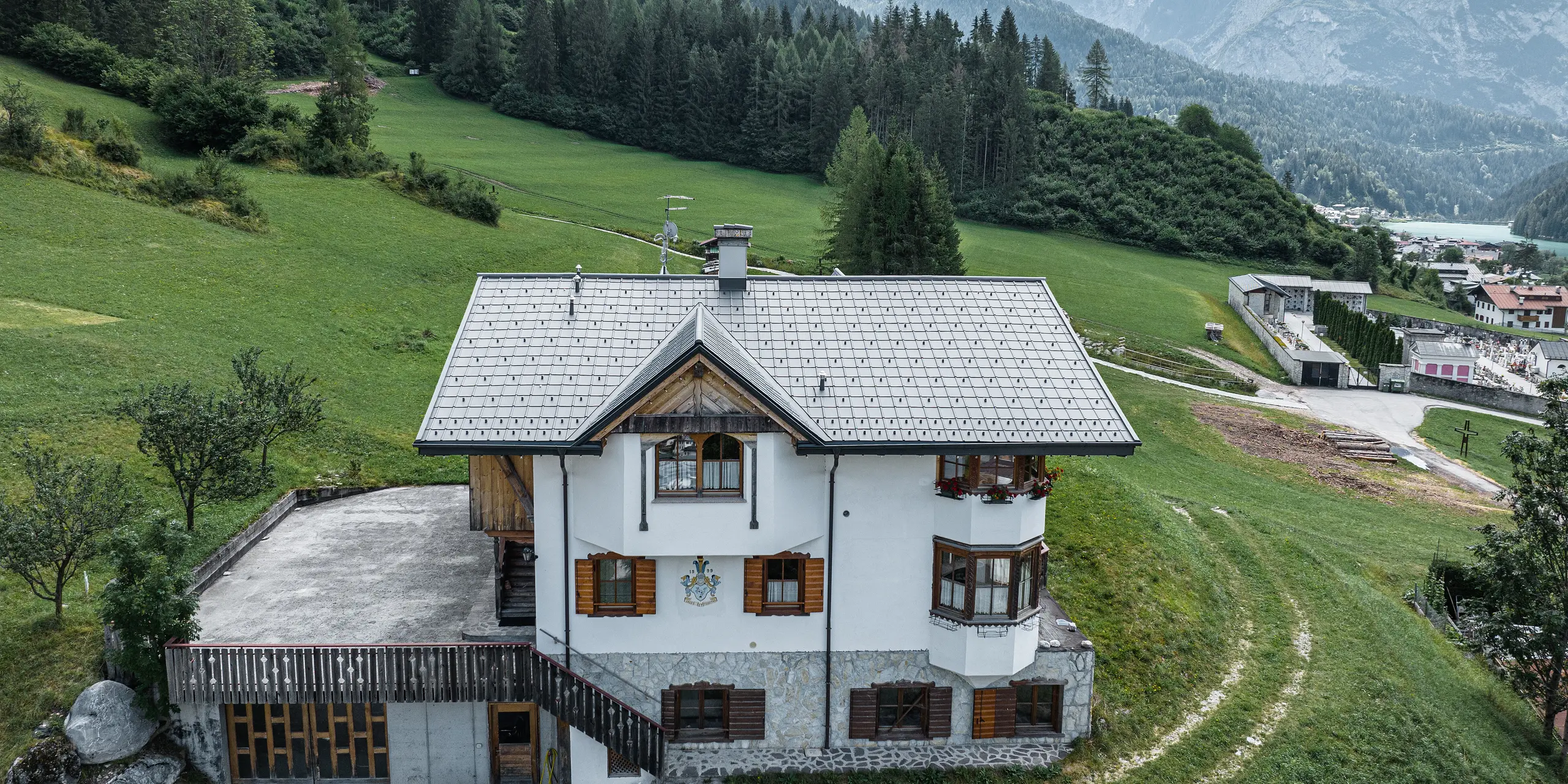 Fugleperspektiv av inngangen til den tradisjonelle fjellhytta i Auronzo di Cadore etter takfornyelsen med PREFA aluminiumsplater i P.10 lys grå. Bygningen med sin karakteristiske alpine sjarm og moderne blikktak føyer seg harmonisk inn i det frodige grønne landskapet og det imponerende fjellpanoramaet i bakgrunnen, og viser en vellykket kombinasjon av funksjonalitet og regional arkitektur.