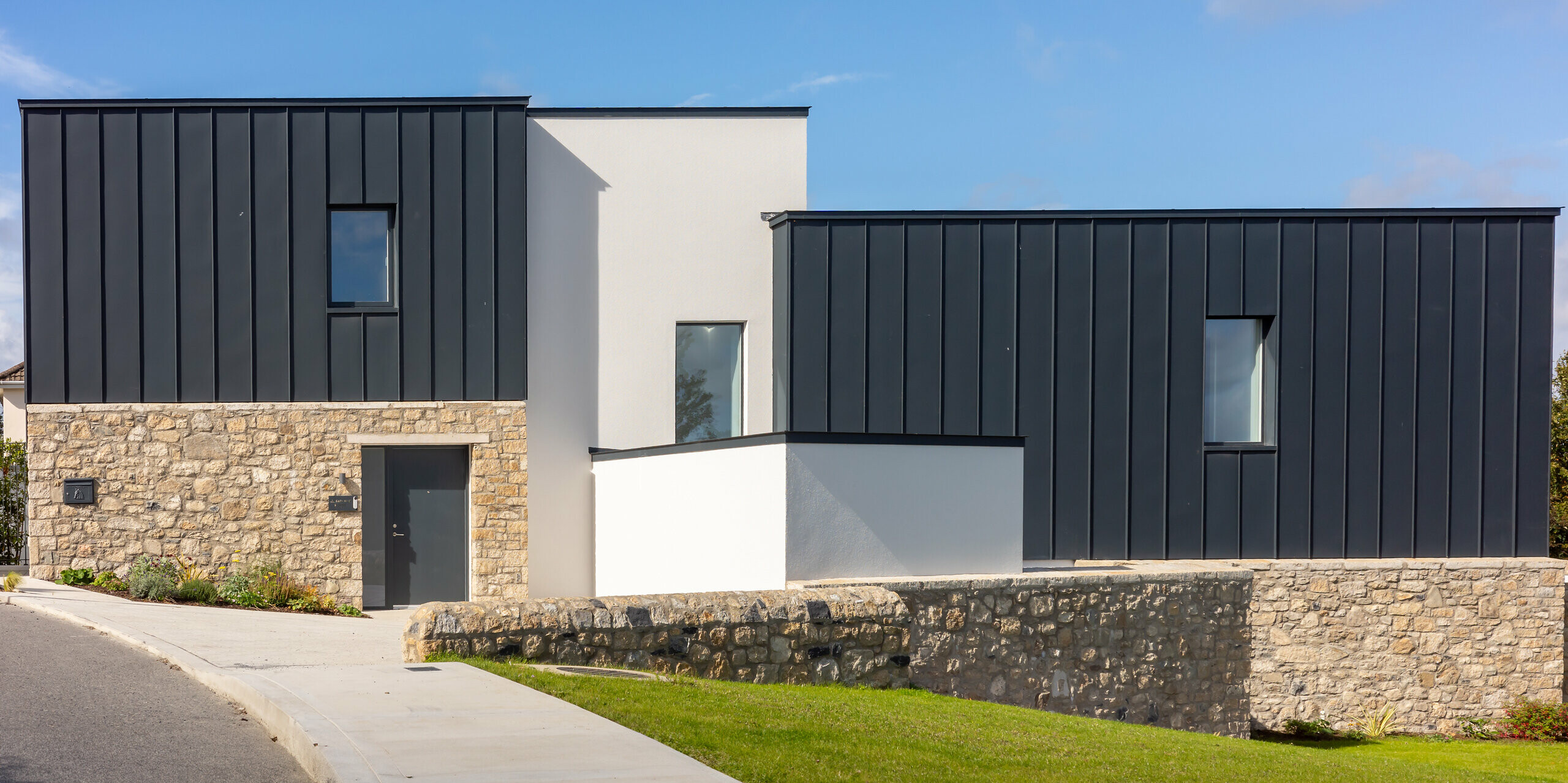 Modernes Wohnhaus in Watson Place mit klaren Linien, Natursteinmauer und markanter PREFALZ Dach‑ und Fassadenverkleidung in P.10 Anthrazit. Die glatten Aluminiumflächen setzen elegante Kontraste zu Stein und Putz und bieten langlebigen Schutz vor Witterung. Das Zusammenspiel der Materialien verleiht dem Gebäude eine hochwertige, zeitlose Erscheinung.