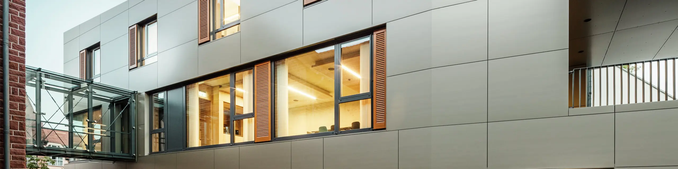Moderne Fassade des Rathauses in Alzenau, Deutschland, mit PREFA-Aluminiumverbundplatten in Bronze, kombiniert mit Fensterläden und begrünten Flächen. Die hochwertige Aluminiumverkleidung sorgt für ein elegantes und langlebiges Erscheinungsbild und ist ideal für nachhaltige Bauprojekte. Die Seite ist hier gut sichtbar.