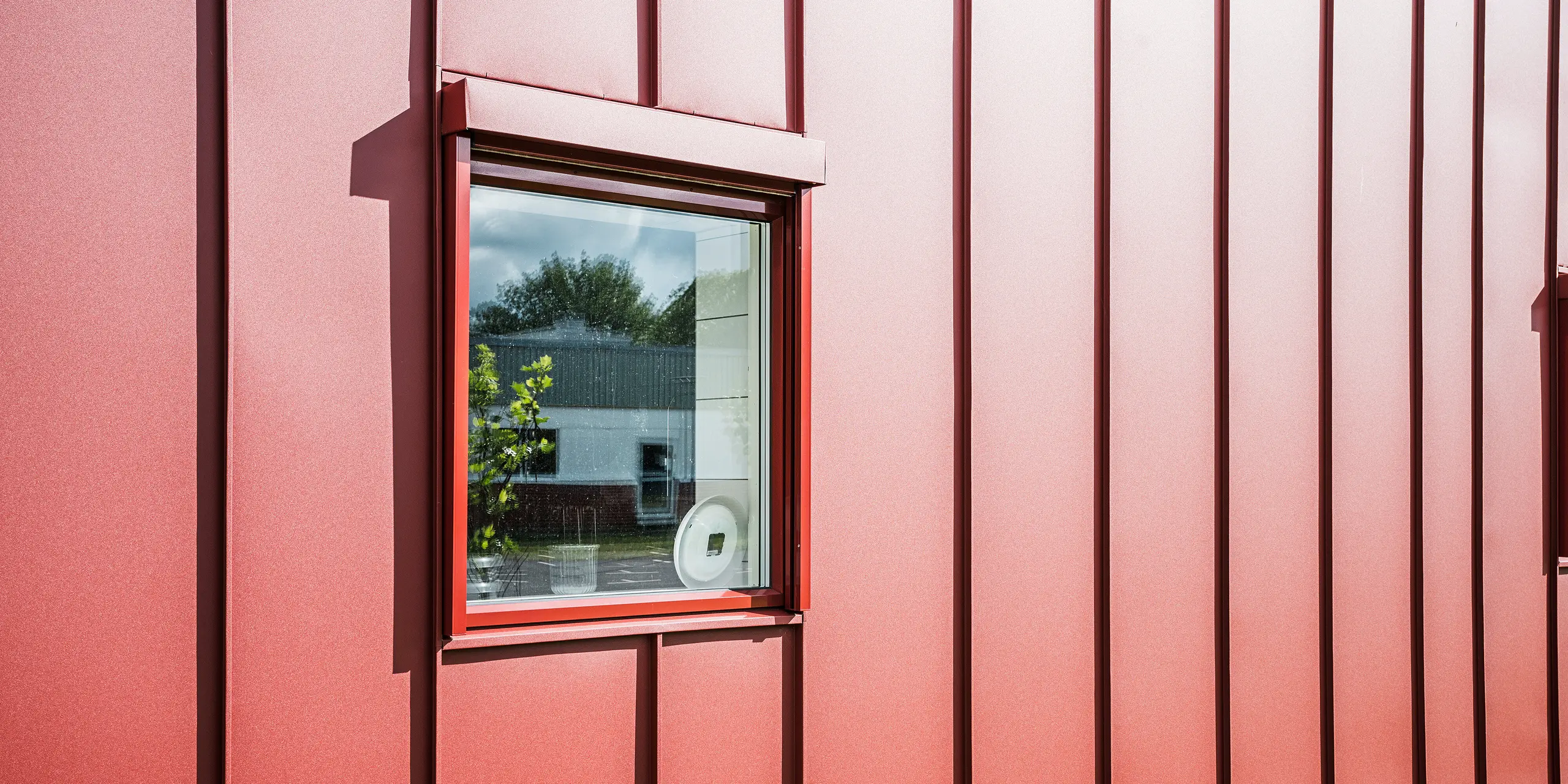 Nahaufnahme eines Fensters der Schulkantine Gröna Hjärtat in Broby, Schweden, zeigt die detailreiche PREFALZ Fassade in Oxydrot. Die präzise Verarbeitung der Stehfalzdeckung aus Aluminium unterstreicht die hohe Qualität und Langlebigkeit der Gebäudehülle. Das Fenster ist harmonisch in die Fassade integriert und reflektiert die Umgebung, was die moderne und klare Architektur des Baukörpers betont. Das ansprechende Design macht das Bauwerk zu einem Beispiel moderner und nachhaltiger Bauweise.