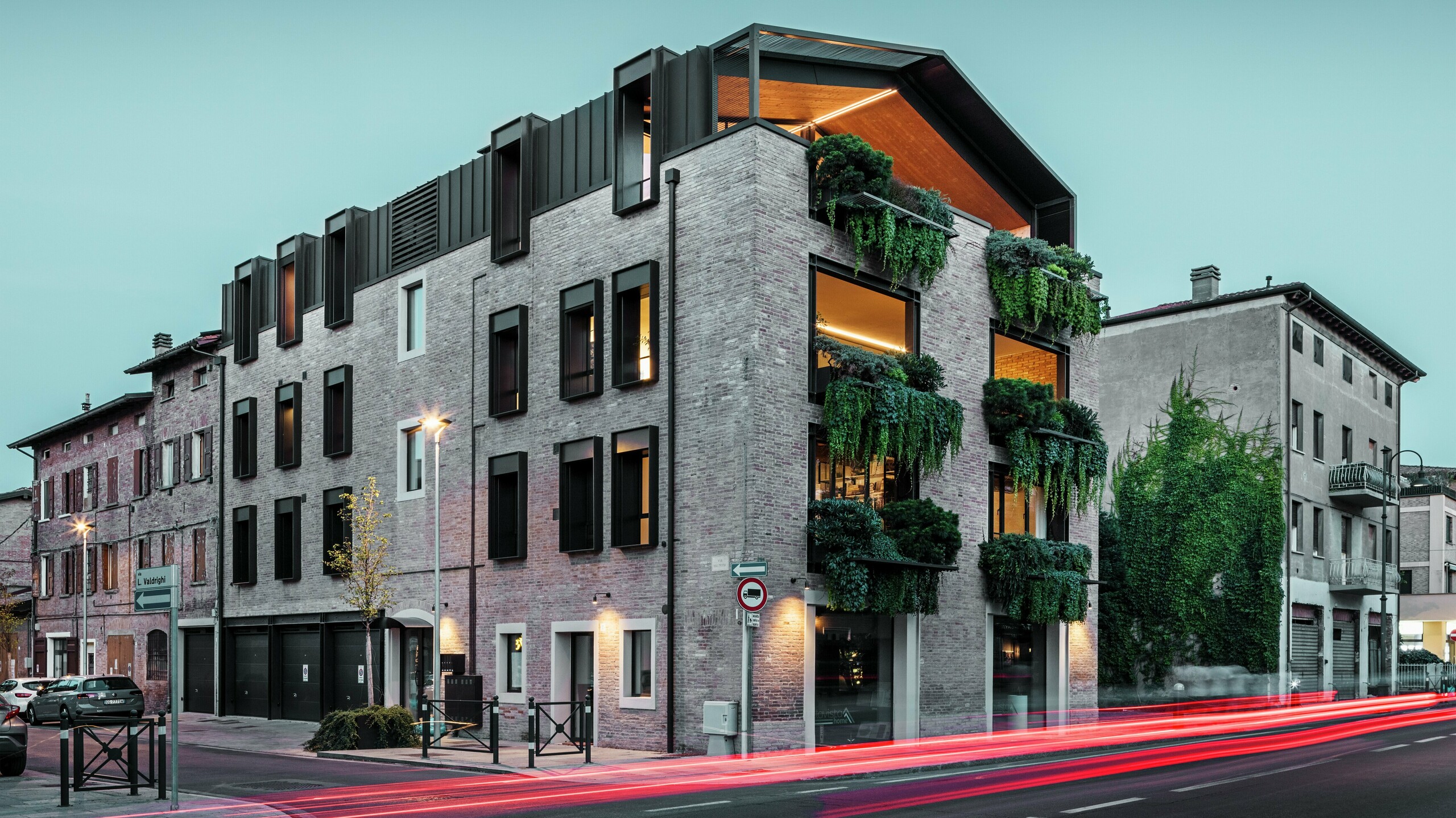 Nachhaltiges Wohn- und Geschäftshaus in Italien mit PREFALZ Aluminiumdach in P.10 Nussbraun, begrünter Backsteinfassade und moderner Architektur bei Abendstimmung mit leuchtenden Straßenlaternen.