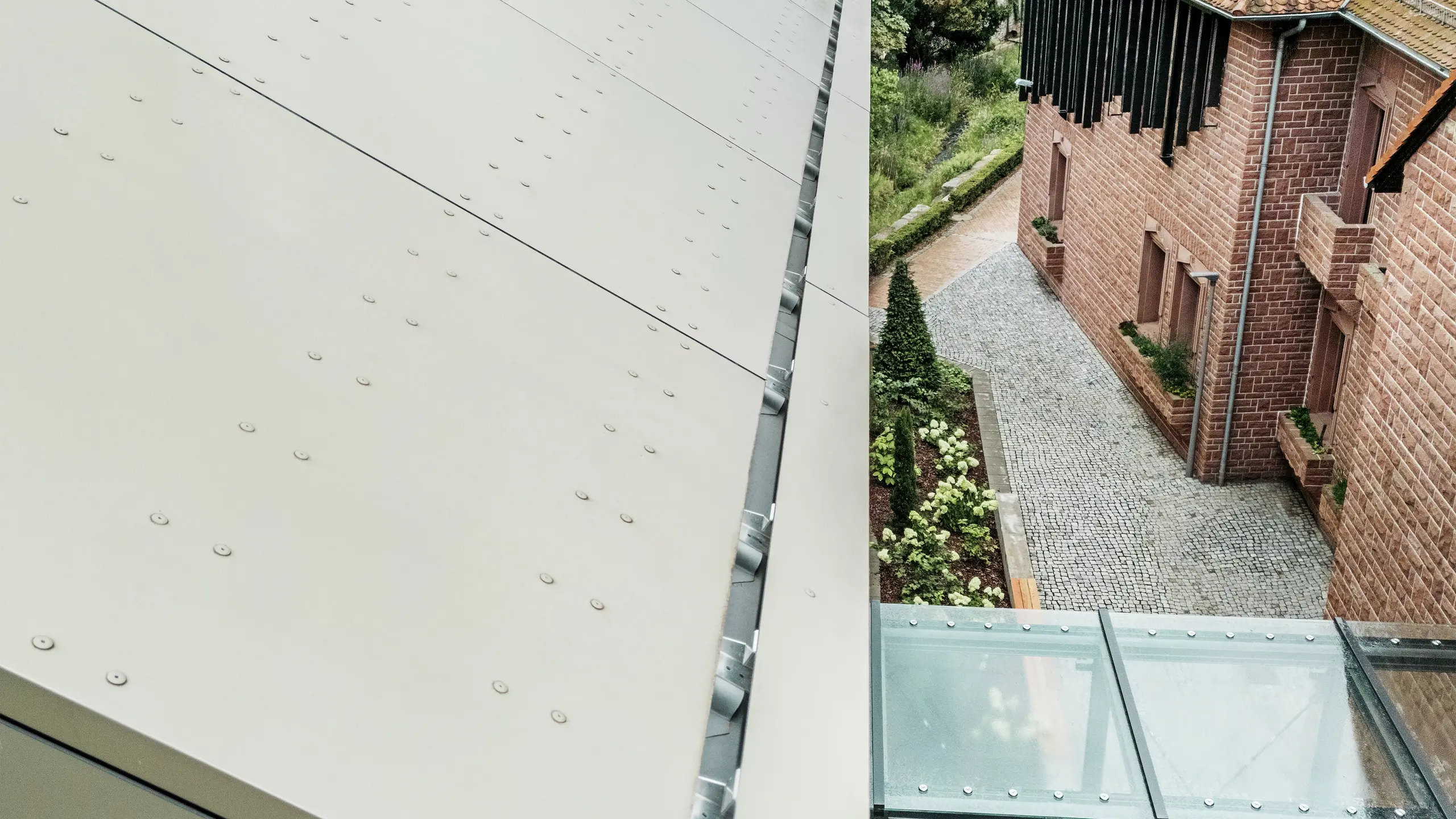 Detailansicht des Daches des Rathauses in Alzenau, das mit PREFA-Aluminiumverbundplatten in einem sanften Bronzeton gedeckt ist. Die Platten sind präzise positioniert und mit sichtbaren Befestigungselementen versehen, was der Fassade ein modernes, industrielles Aussehen verleiht. Im Vordergrund ist der gläserne Verbindungsgang zu sehen, der das Rathaus mit dem Nachbargebäude verbindet.