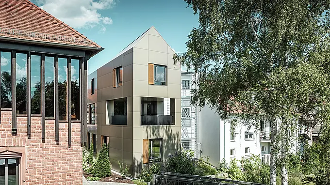 Das Bild zeigt das moderne Rathaus in Alzenau, dessen Fassade aus Aluminium-Verbundplatten von PREFA in Bronze besteht. Der spitze Dachbereich und die klar strukturierten Fenster mit Holzjalousien verleihen dem Gebäude eine elegante und zeitgenössische Ästhetik. Im Vordergrund ist ein grüner Bereich mit einem gepflasterten Weg und Bäumen zu sehen, der das urbane Design des Gebäudes harmonisch ergänzt.