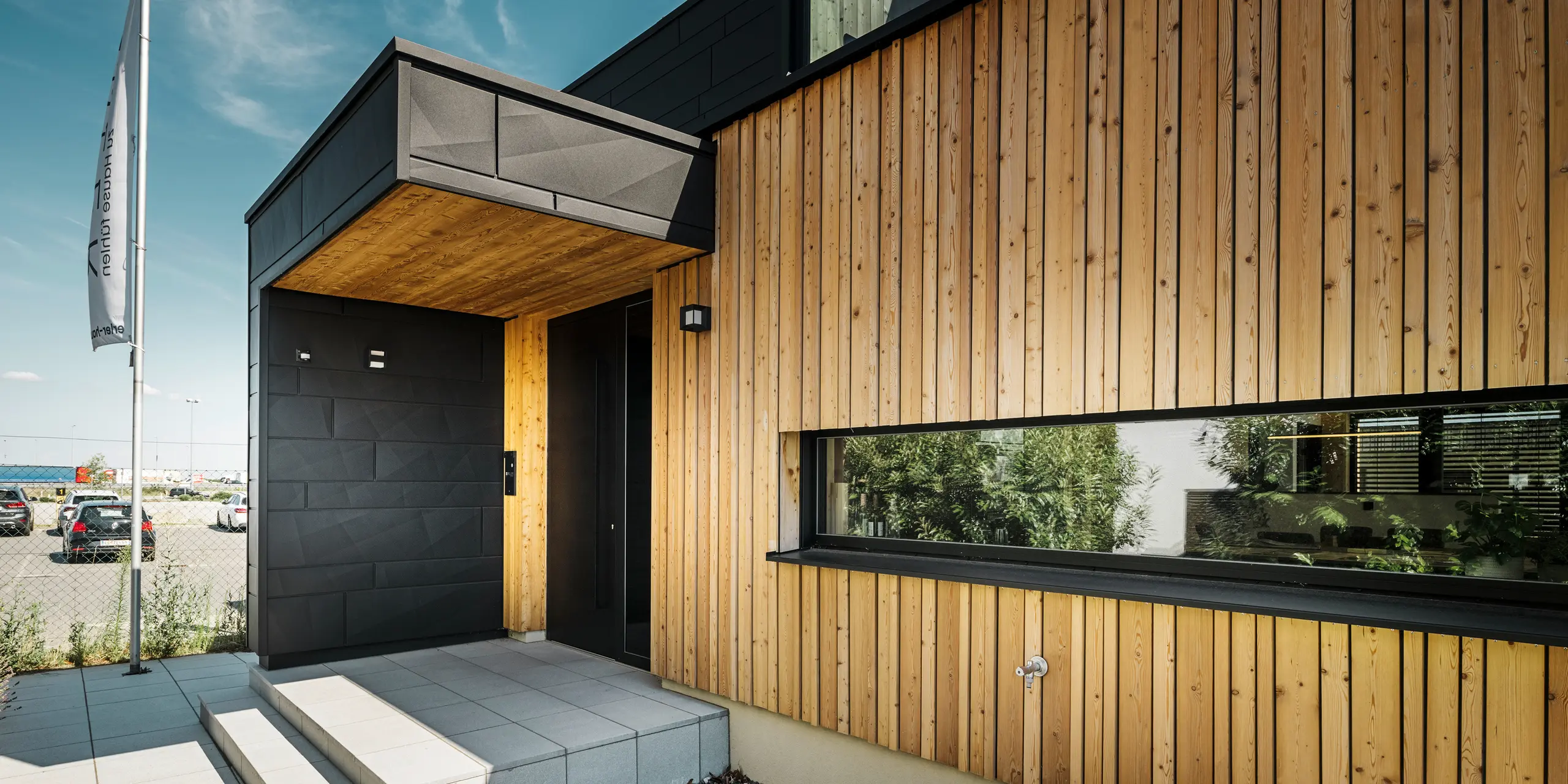 Sideinngangen til utstillingshuset Autark i ferdighussenteret Blaue Lagune i Vösendorf, med en moderne fasade av naturlig tre og svarte PREFA Siding.X-paneler. Bildet viser den elegante kombinasjonen av trekledning og aluminium i høykvalitetsbelegget P.10 Black.
