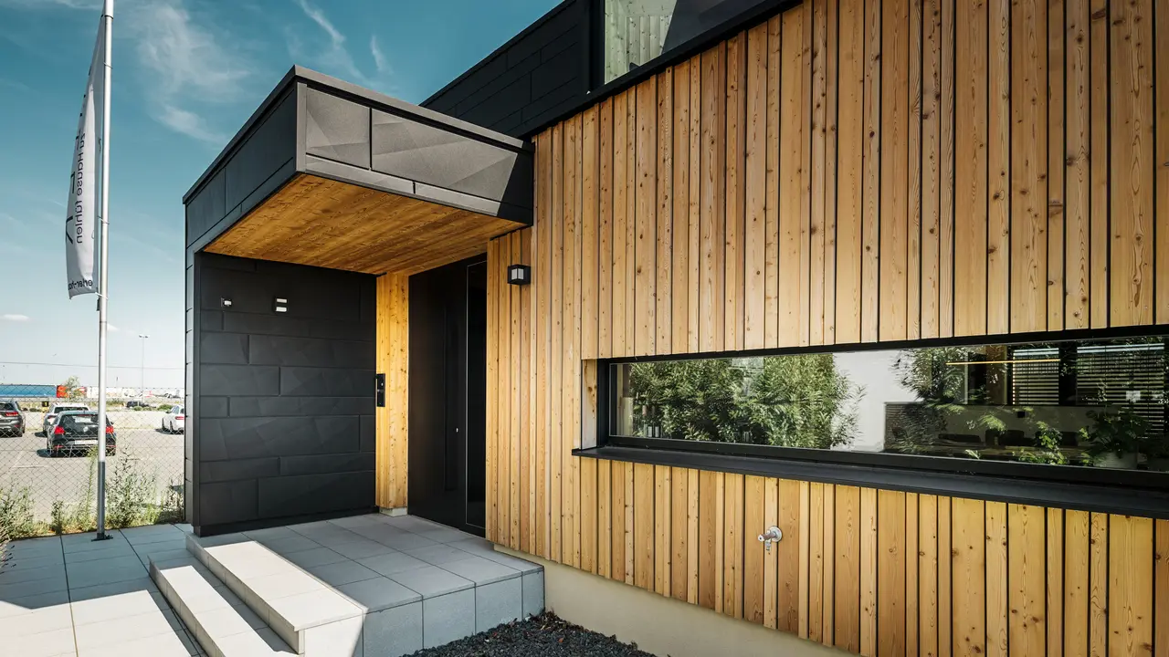 Sideinngangen til utstillingshuset Autark i ferdighussenteret Blaue Lagune i Vösendorf, med en moderne fasade av naturlig tre og svarte PREFA Siding.X-paneler. Bildet viser den elegante kombinasjonen av trekledning og aluminium i høykvalitetsbelegget P.10 Black.