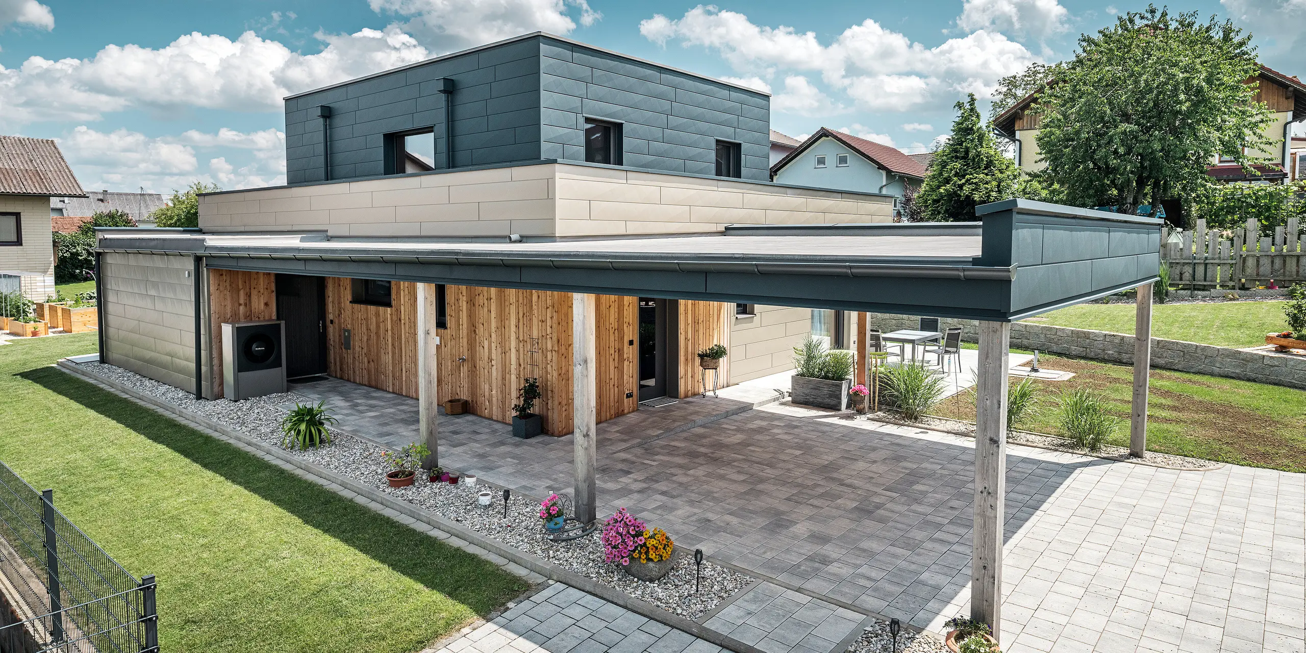 Luftfoto av oppkjørselen til en moderne enebolig i Wippenham, Østerrike, med en tofarget PREFA aluminiumsfasade i bronse og antrasitt. Huset viser den stilfulle integreringen av Siding.X fasadepaneler og trebekledning. Den overbygde carporten, som er sømløst integrert i husets arkitektur, er tydelig synlig. Den velstelte hagen og terrasseområdet understreker bygningens moderne og funksjonelle design.