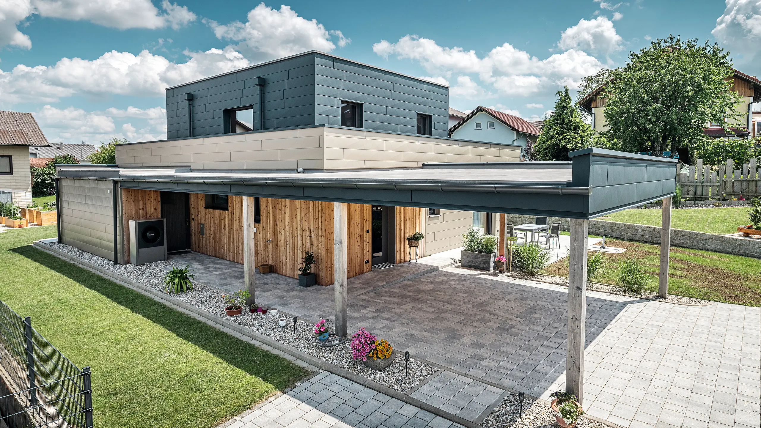 Luftfoto av oppkjørselen til en moderne enebolig i Wippenham, Østerrike, med en tofarget PREFA aluminiumsfasade i bronse og antrasitt. Huset viser den stilfulle integreringen av Siding.X fasadepaneler og trebekledning. Den overbygde carporten, som er sømløst integrert i husets arkitektur, er tydelig synlig. Den velstelte hagen og terrasseområdet understreker bygningens moderne og funksjonelle design.