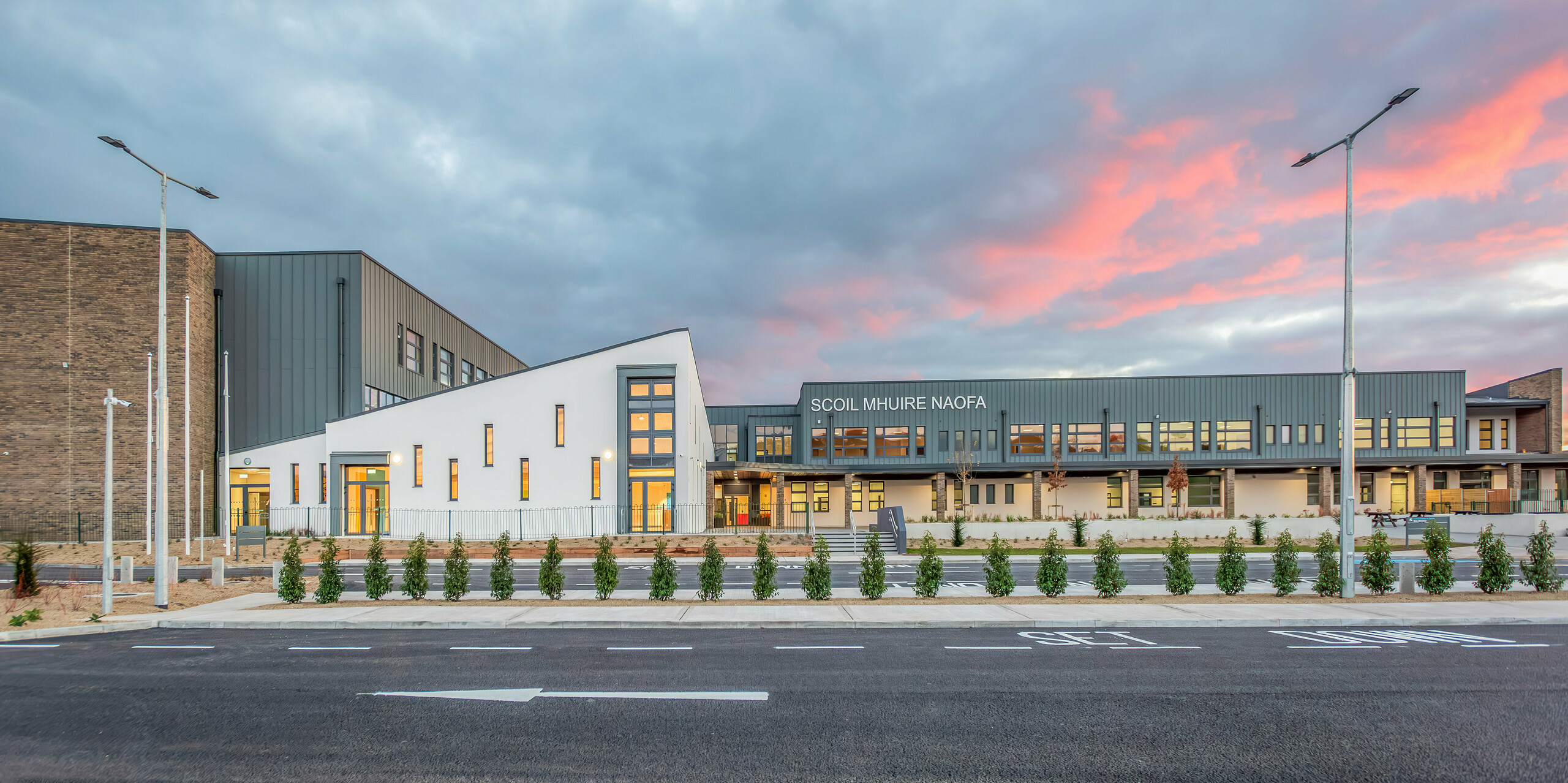 Frontansicht der neuen Scoil Mhuire Naofa in Carrigtwohill, Irland, bei Sonnenuntergang. Die hellgraue PREFALZ Aluminiumfassade kombiniert mit modernen Ziegeldetails und klaren Linien schafft eine harmonische und langlebige Architektur. Der großzügige Außenbereich unterstreicht die einladende Atmosphäre der Schule.