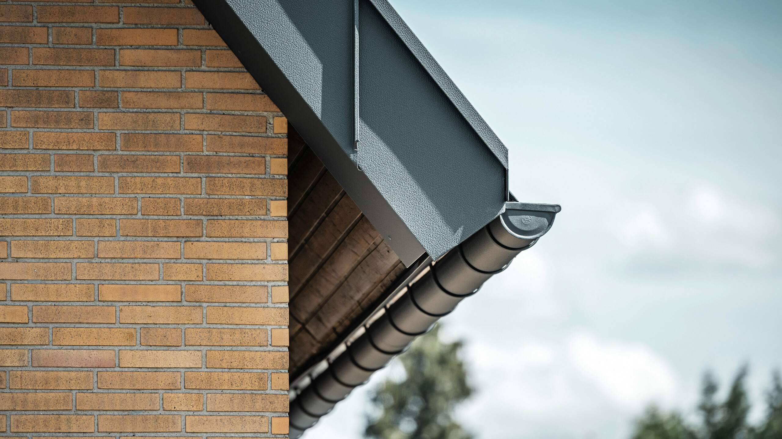 Detaljbilde av et antrasittfarget takrenne-system fra PREFA. Takrennen skiller seg tydelig fra de varme fargene på murveggen og understreker den presise bearbeidelsen og kvaliteten på materialene som er brukt.