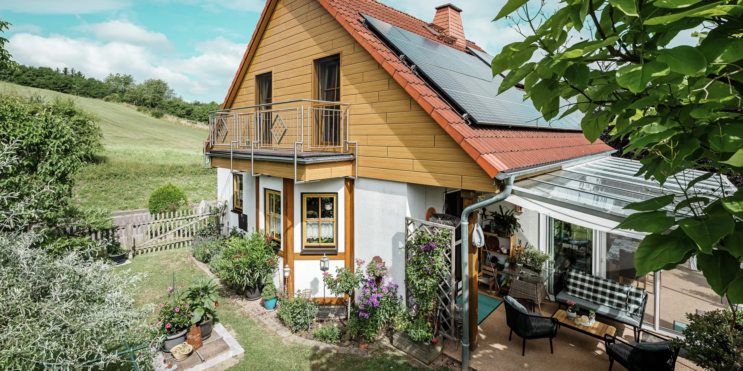 Dette bildet viser en sjarmerende enebolig i Blieskastel, der en nyrenovert gavl med PREFA Siding i natureik er særlig iøynefallende. Den varme treoverflaten i slitesterkt aluminium passer svært godt sammen med det røde tegltaket og den romslige vinterhagen. Omgitt av frodig grønt og blomsterbed anlagt med omtanke, fremstår helheten som idyllisk og levende. Den robuste renoveringen av fasader med PREFA gir ikke bare huset et naturlig uttrykk, men imponerer også med god bestandighet mot vær, stabil farge og en tilnærmet vedlikeholdsfri levetid.