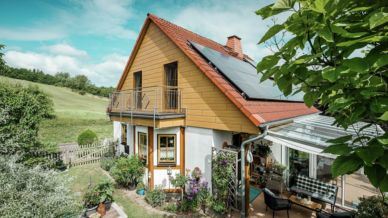Dette bildet viser en sjarmerende enebolig i Blieskastel, der en nyrenovert gavl med PREFA Siding i natureik er særlig iøynefallende. Den varme treoverflaten i slitesterkt aluminium passer svært godt sammen med det røde tegltaket og den romslige vinterhagen. Omgitt av frodig grønt og blomsterbed anlagt med omtanke, fremstår helheten som idyllisk og levende. Den robuste renoveringen av fasader med PREFA gir ikke bare huset et naturlig uttrykk, men imponerer også med god bestandighet mot vær, stabil farge og en tilnærmet vedlikeholdsfri levetid.