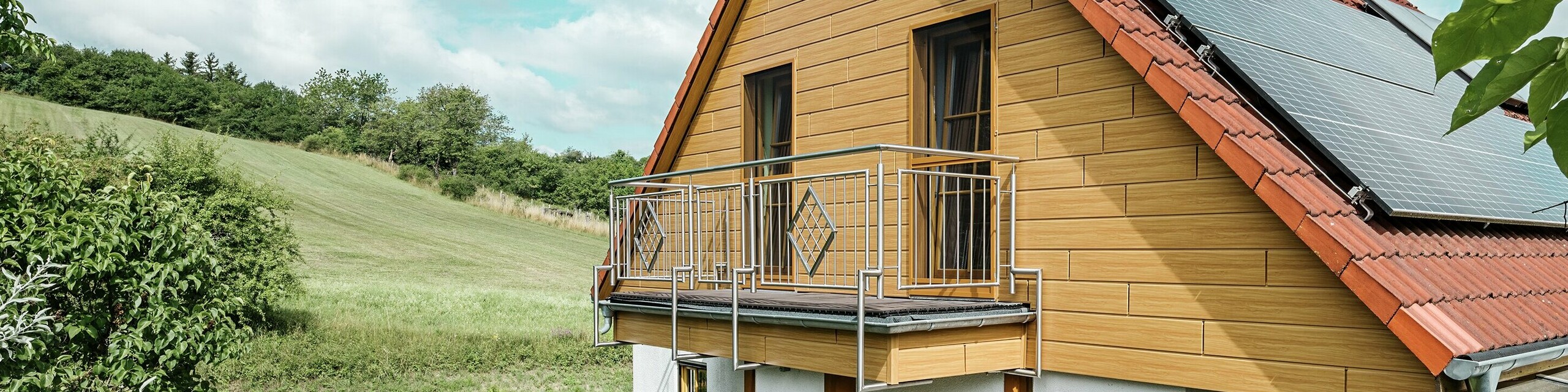 Dette bildet viser en sjarmerende enebolig i Blieskastel, der en nyrenovert gavl med PREFA Siding i natureik er særlig iøynefallende. Den varme treoverflaten i slitesterkt aluminium passer svært godt sammen med det røde tegltaket og den romslige vinterhagen. Omgitt av frodig grønt og blomsterbed anlagt med omtanke, fremstår helheten som idyllisk og levende. Den robuste renoveringen av fasader med PREFA gir ikke bare huset et naturlig uttrykk, men imponerer også med god bestandighet mot vær, stabil farge og en tilnærmet vedlikeholdsfri levetid.