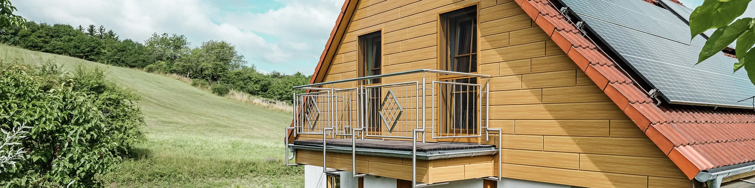 Dette bildet viser en sjarmerende enebolig i Blieskastel, der en nyrenovert gavl med PREFA Siding i natureik er særlig iøynefallende. Den varme treoverflaten i slitesterkt aluminium passer svært godt sammen med det røde tegltaket og den romslige vinterhagen. Omgitt av frodig grønt og blomsterbed anlagt med omtanke, fremstår helheten som idyllisk og levende. Den robuste renoveringen av fasader med PREFA gir ikke bare huset et naturlig uttrykk, men imponerer også med god bestandighet mot vær, stabil farge og en tilnærmet vedlikeholdsfri levetid.