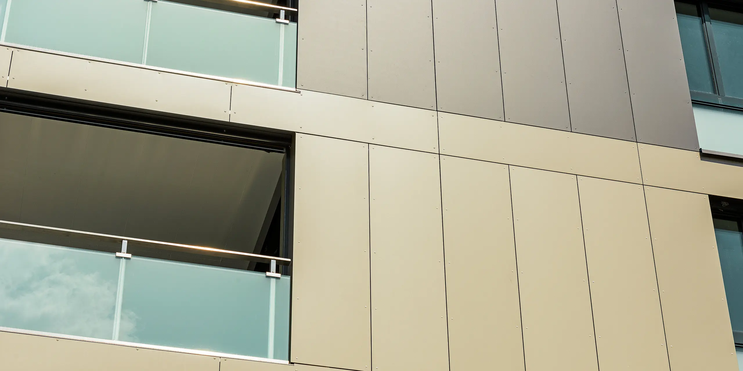 Nahaufnahme der Aluminiumfassade an der kubischen Wohnanlage in Murten (CH). Die Aluminium Verbundplatten von PREFA garantieren eine hohe Widerstandsfähigkeit und Langlebigkeit. Die harmonische Farbgestaltung in Bronze und Schwarzgrau fügt sich perfekt in die Umgebung ein und schützt das Gebäude effektiv vor äußeren Einflüssen wie Wind und Wetter. An den Bildrändern sind Balkone mit Glasverkleidungen zu sehen.