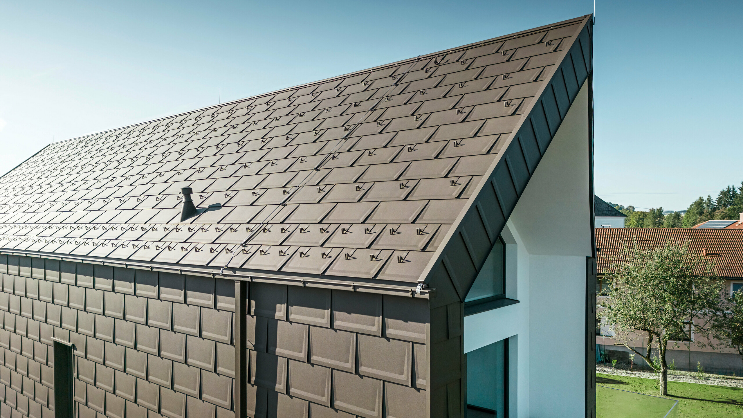 Perspektivisk visning av tak og fasade på en moderne enebolig i Neukirchen, Østerrike. PREFA R.16 takplate i brunfarge P.10 gir en estetisk og funksjonell takbelegg som passer perfekt til husets arkitektoniske utforming. Den skarpe mønen på taket og den presise plasseringen av takplatene viser kvaliteten til PREFA. De småformat aluminiumproduktene passer perfekt inn i den hvitpussede fasaden og det grønne landskapet rundt.