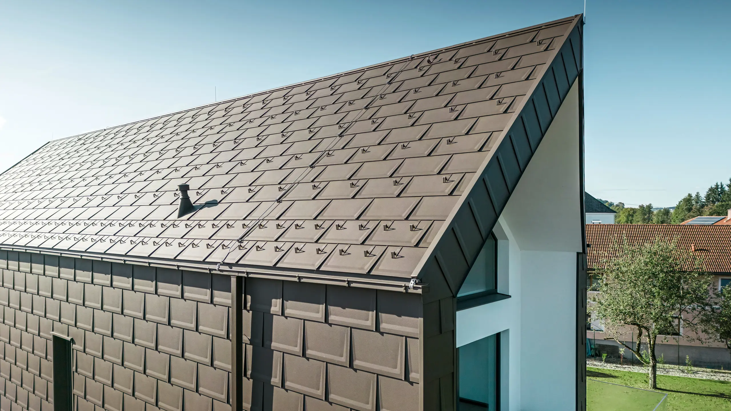 Perspektivisk visning av tak og fasade på en moderne enebolig i Neukirchen, Østerrike. PREFA R.16 takplate i brunfarge P.10 gir en estetisk og funksjonell takbelegg som passer perfekt til husets arkitektoniske utforming. Den skarpe mønen på taket og den presise plasseringen av takplatene viser kvaliteten til PREFA. De småformat aluminiumproduktene passer perfekt inn i den hvitpussede fasaden og det grønne landskapet rundt.