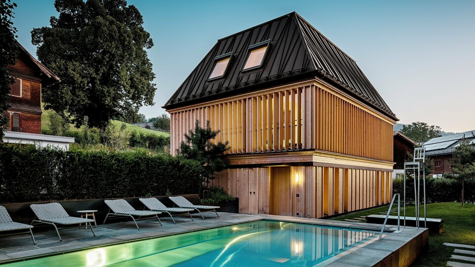 Das „Badehaus“ des Hotel Hirschen in Schwarzenberg in der Dämmerung, mit Blick auf den beleuchteten Außenpool im Vordergrund. Das Gebäude beeindruckt durch sein modernes Walmdach aus PREFALZ in der Farbe P.10 Nussbraun mit präzise ausgeführten, teils schräg verlaufenden Stehfalzen. Zwei Dachfenster sorgen für Tageslicht im Inneren. Die Fassade besteht aus vertikalen, geschoßhohen Holzlamellen, die dem Baukörper Struktur und Leichtigkeit verleihen. Im unteren Bereich dominiert naturbelassenes Holz. Umgeben von gepflegtem Grün und eingebettet in die Hügellandschaft des Bregenzerwaldes entsteht eine harmonische Verbindung aus Architektur, Natur und Erholung.