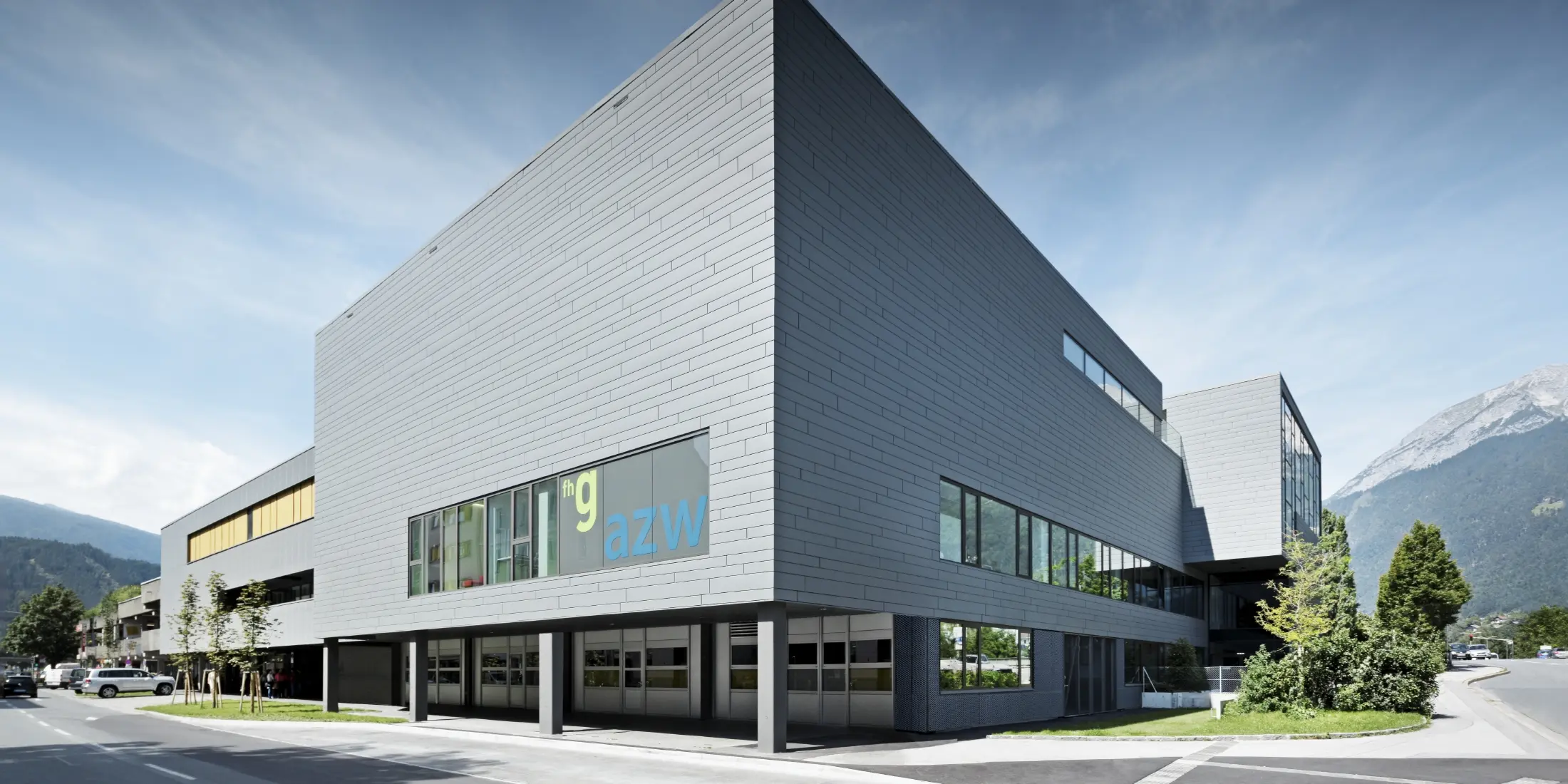 Modernes Ausbildungszentrum mit klarer, kubischer Architektur und hellgrauer Aluminiumfassade. Das Gebäude hat große Fensterflächen und überhängende Baukörper. Auf einer Glasfront ist die Beschriftung „fhg azw“ zu sehen. Im Hintergrund erheben sich Berge unter einem leicht bewölkten Himmel, während im Vordergrund eine gepflegte Straße mit Grünflächen verläuft.