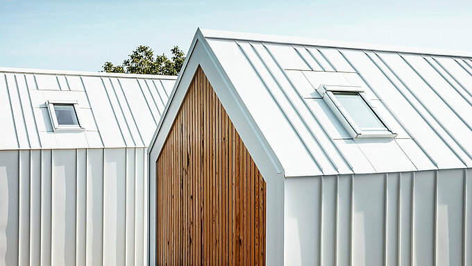 Detailansicht der weißen PREFALZ Dach- und Fassadenverkleidung der Tiny Häuser 'Pri Momi' in Kančevci, Slowenien. Die Gebäude sind in einer modernen Kombination aus hochwertigem Aluminium in P.10 Reinweiß und naturbelassenem Holz gestaltet. Das Bild zeigt die präzise ausgeführten Falze und den eleganten Übergang vom Dach zur Fassade, der ohne sichtbare Unterbrechung der vertikalen Stehfalze realisiert wurde. Die harmonische Materialwahl unterstreicht das minimalistische Design der exklusiven Gästeunterkunft.