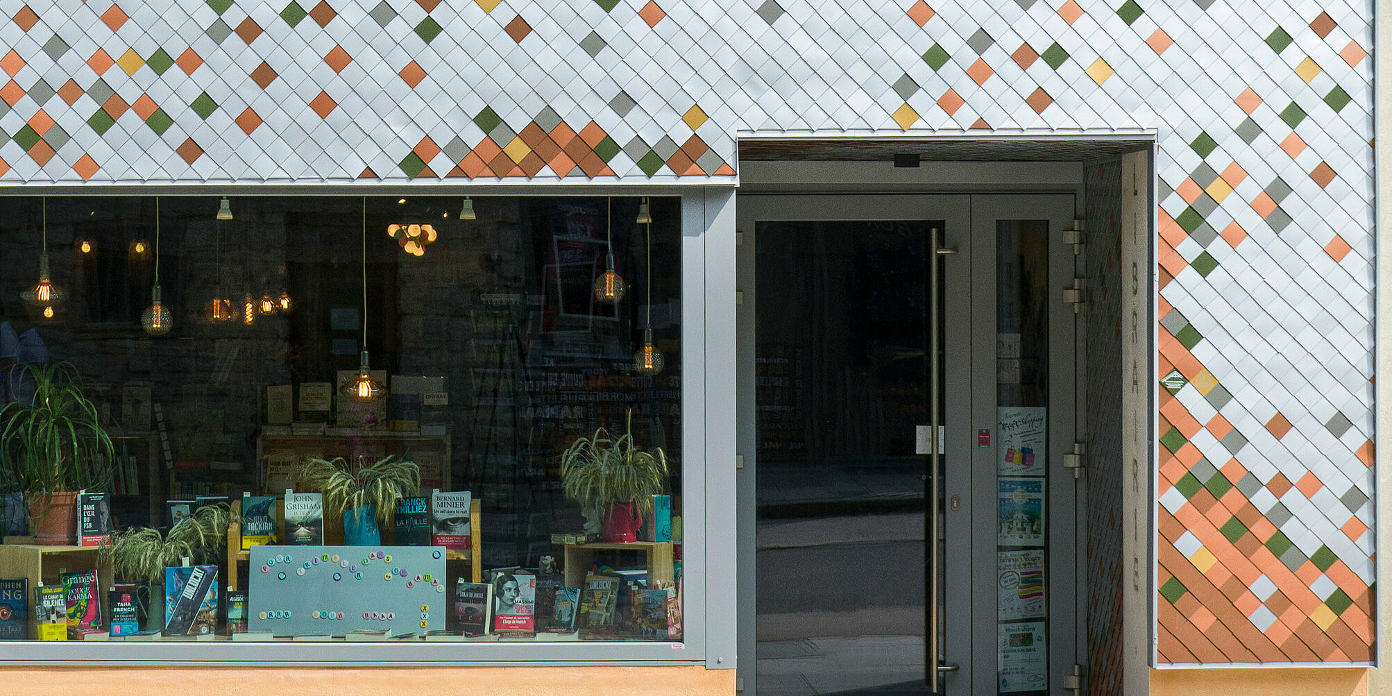 Frontansicht der bunten Fassade einer Buchhandlung in der kleinen französischen Gemeinde Les Rousses. Das Gebäude kombiniert ein leuchtend orangefarbenes Obergeschoss mit dekorativen Elementen und ein unteres Geschoss mit einer mosaikartigen Fassade aus Aluminium in verschiedenen Farben. Ein großes Schaufenster im Erdgeschoss präsentiert Bücher und Pflanzen neben der Eingangstür. Eine schmale Dachrinne verläuft an der Seite des Gebäudes, und die Nummer 55 ist neben der Tür auf einem grünen Schild zu sehen.