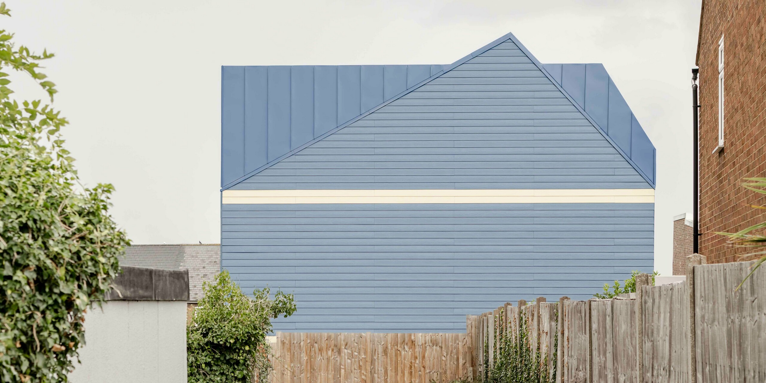 Die Aufnahme zeigt die markante Stirnseite eines Goldsmith‑Mews‑Reihenhauses, verkleidet mit taubenblauem PREFA FALZONAL Aluminium. Die klaren Linien der vertikalen und horizontalen Falzstrukturen erzeugen ein kraftvolles, geometrisches Fassadenbild, das sich deutlich von der umgebenden Bebauung abhebt. Der Kontrast zu den hellen unteren Fassadenbereichen verstärkt die moderne, präzise Gestaltung. Das langlebige, farbstabile PREFA Aluminium bietet hohen Witterungsschutz und sorgt für eine dauerhaft hochwertige Optik, die dem Ensemble seine einprägsame Identität verleiht.