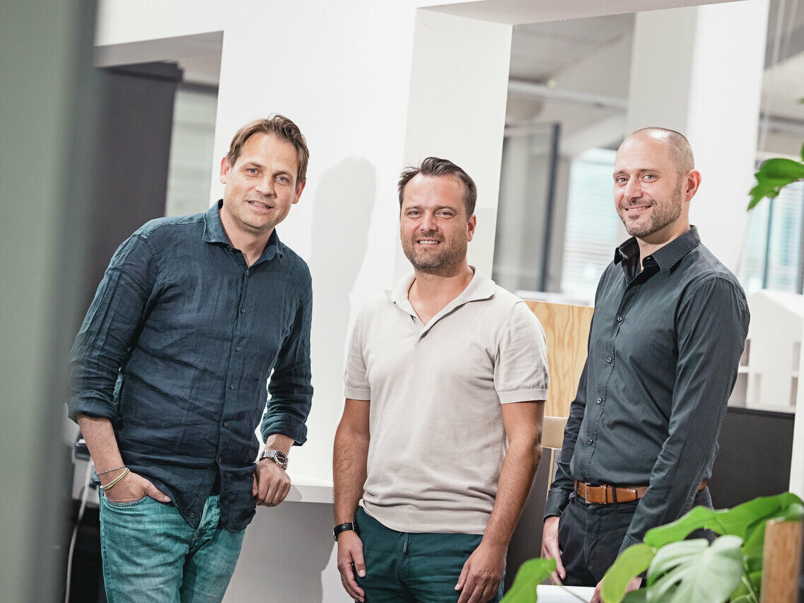 Das Architektenteam des Architektur- und Designstudios CBA stehen in einem hellen Büro. Die Kleidung ist leger und professionell, sie sind gekleidet in Hemden und Poloshirts. Im Hintergrund ist ein modernes Büroumfeld mit weißen Säulen und Zimmerpflanzen zu sehen.