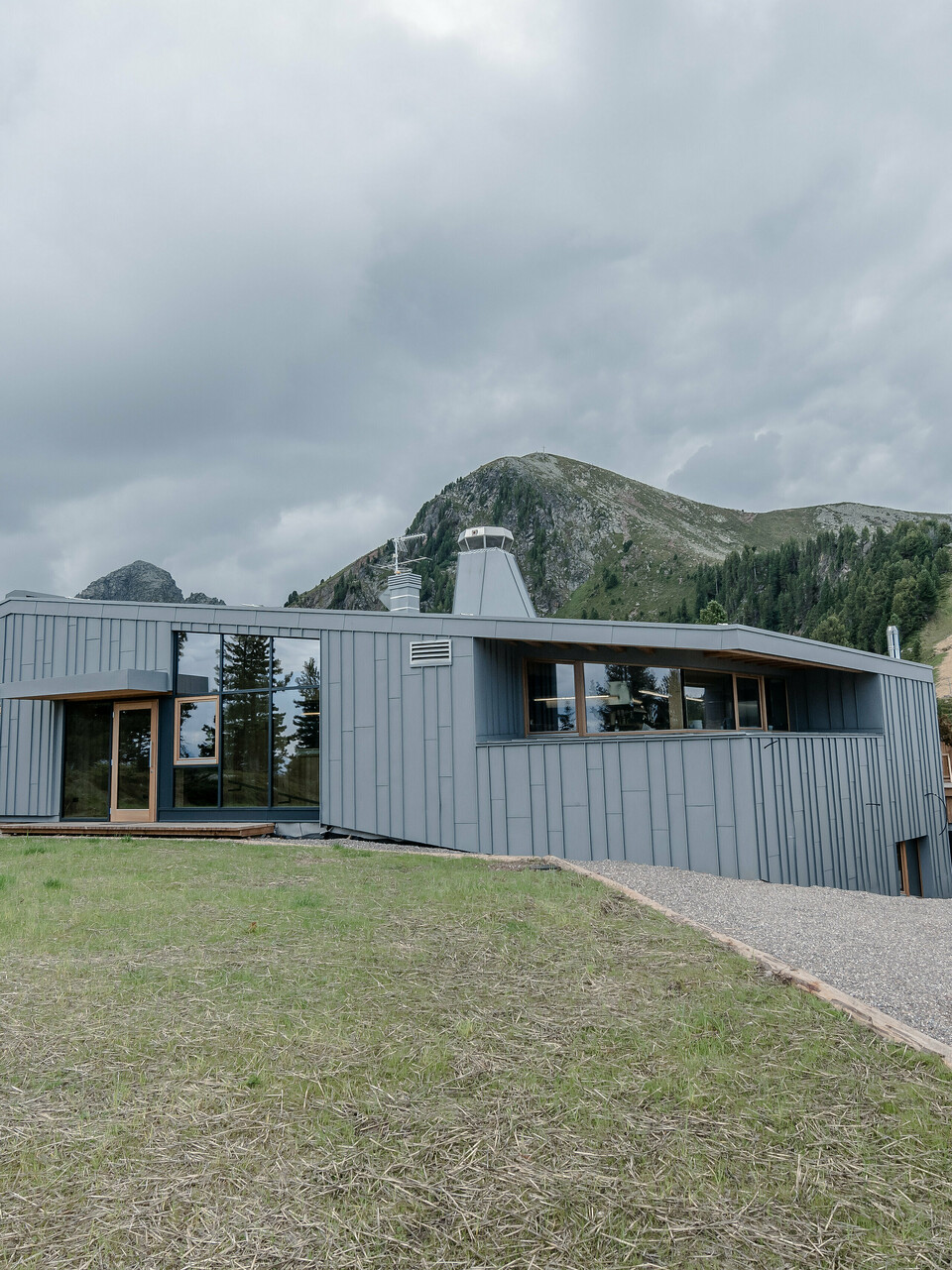 Das Ristorante Busabelle, eingebettet in eine idyllische Berglandschaft, ist von einer Winkelstehfalz Spiegeldeckung aus hellgrauem PREFALZ ummantelt, die die moderne Architektur hervorhebt und eine harmonische Verbindung zur natürlichen Umgebung schafft. Der Eingangsbereich ist mit großen Glasflächen versehen. Im Hintergrund sind Masten für eine Sesselliftanlage zu erkennen. 