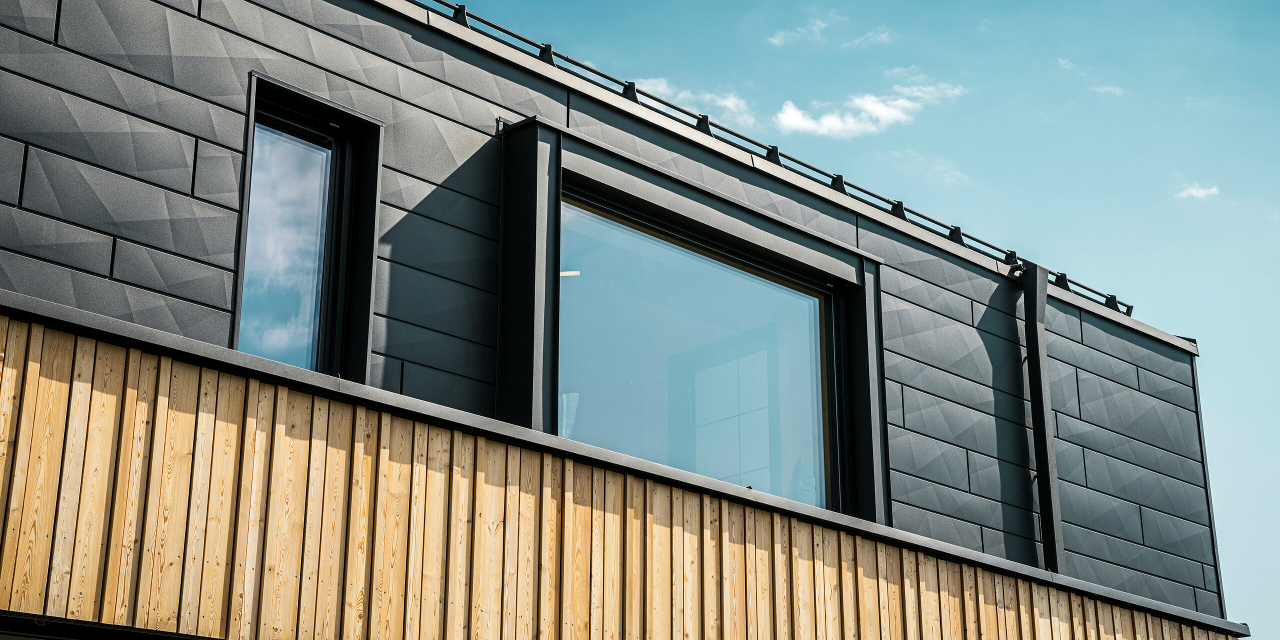 Nahaufnahme des Obergeschosses des Musterhauses Autark im Fertighauszentrum Blaue Lagune, Vösendorf (AT). Die Fassade besteht aus PREFA Siding.X Aluminiumpaneelen in P.10 Schwarz, die durch ihre spezielle Optik mit unterschiedlichen Kantungen auffallen. Darunter befindet sich eine stilvolle Holzverkleidung, die einen harmonischen Kontrast zum dunklen Aluminium bildet. Große Fenster lassen viel Licht in das Gebäudeinnere und betonen das moderne Design.