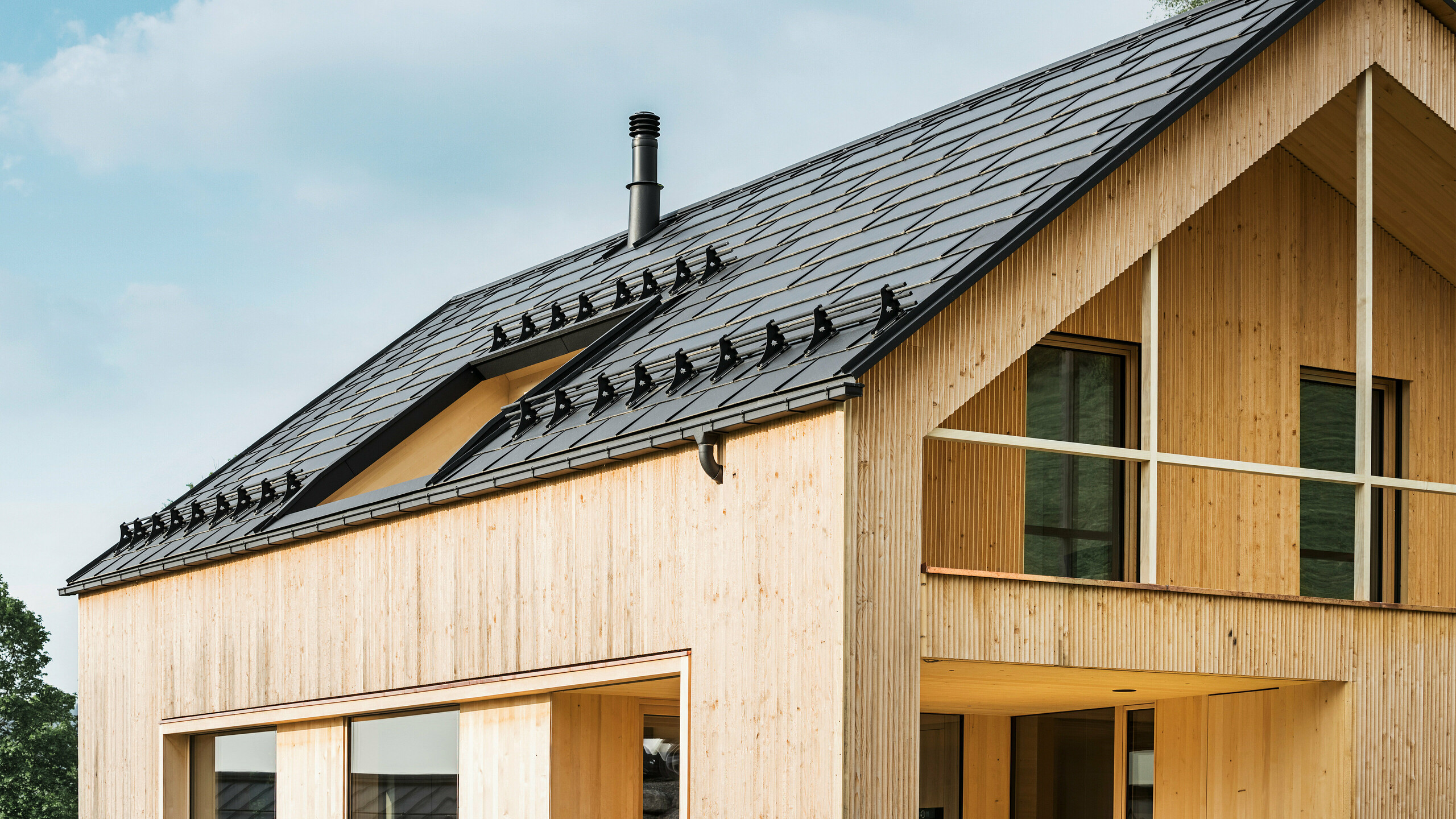 Nærbilde av gavlen på et miljøvennlig hus i Egg, preget av PREFA solcelle takplater i elegant svart, som harmonerer med PREFA takplater R.16. De rene linjene på taket står i kontrast til den naturlige strukturen på trefasaden, mens solcellemodulene er et statement for bærekraftig boligbygging. Den diskrete skorsteinen passer inn i helhetsbildet, som kombinerer miljøbevisst design med landlig estetikk. I tillegg har det innovative økohuset to balkonger som gir utsikt over den fantastiske fjellkulissen i Egg i Vorarlberg.