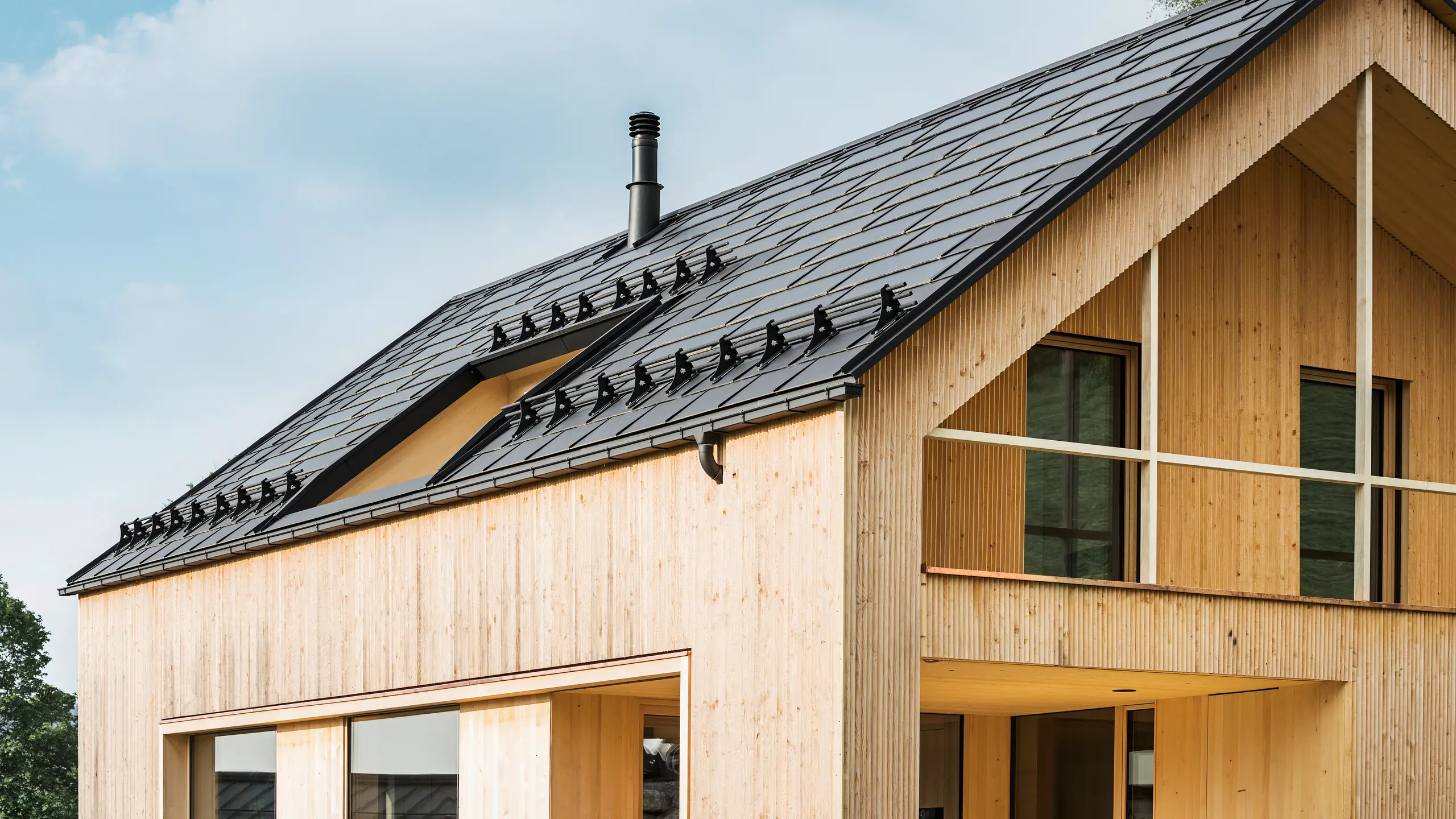 Nærbilde av gavlen på et miljøvennlig hus i Egg, preget av PREFA solcelle takplater i elegant svart, som harmonerer med PREFA takplater R.16. De rene linjene på taket står i kontrast til den naturlige strukturen på trefasaden, mens solcellemodulene er et statement for bærekraftig boligbygging. Den diskrete skorsteinen passer inn i helhetsbildet, som kombinerer miljøbevisst design med landlig estetikk. I tillegg har det innovative økohuset to balkonger som gir utsikt over den fantastiske fjellkulissen i Egg i Vorarlberg.