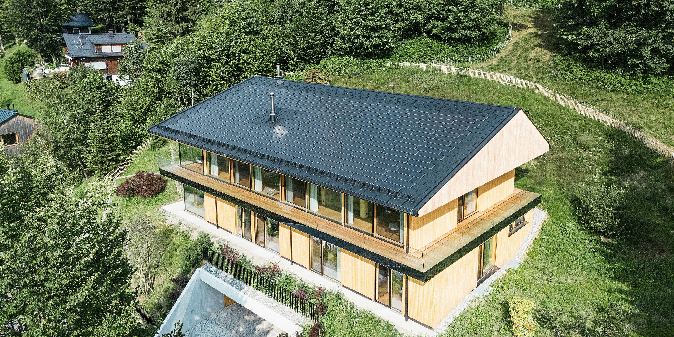 Vogelperspektive auf ein Einfamilienhaus in Egg, Vorarlberg, mit PREFA Solardachplatten und Dachplatten R.16 in P.10 Schwarz. Das moderne Aluminiumdach mit integrierter Photovoltaikanlage sorgt für nachhaltige Energiegewinnung. Die elegante Holzfassade harmoniert perfekt mit der natürlichen Waldlandschaft im Hintergrund. Der robuste Schneeschutz und die Dachentwässerung von PREFA sorgen für Sicherheit und Funktionalität.