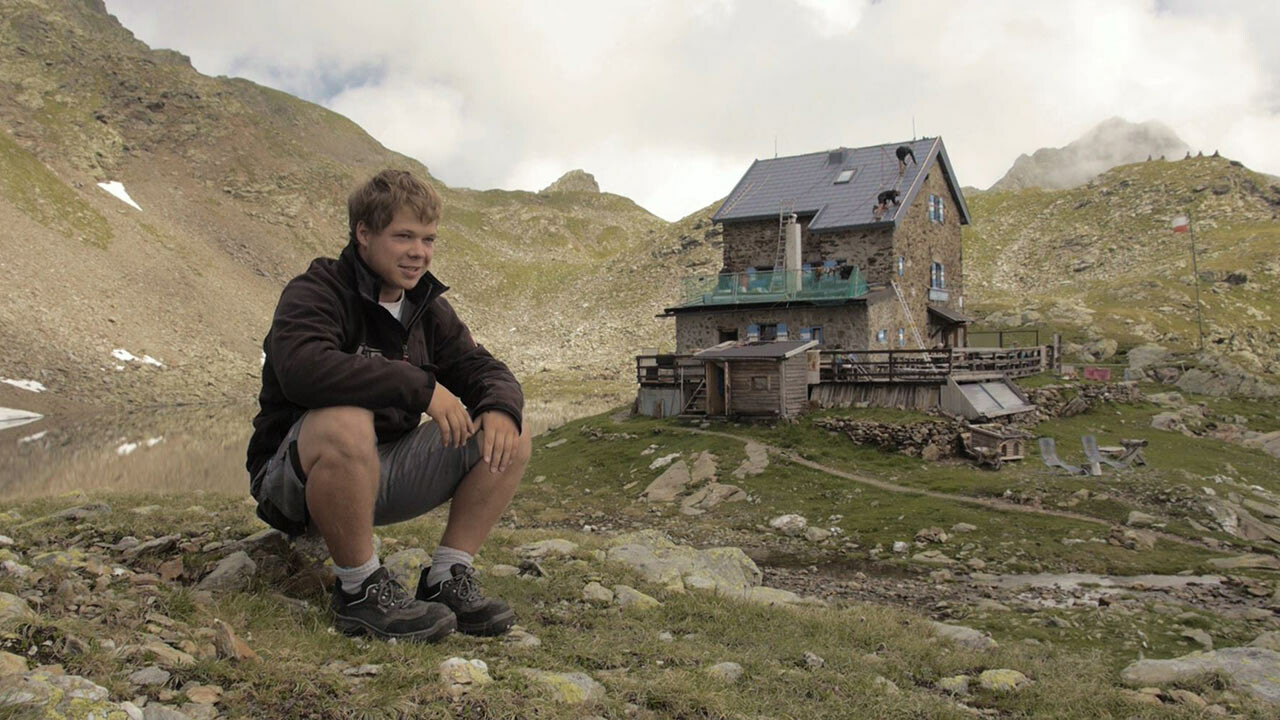 Lærlingen Jakob Schöttl sidder på en sten. I baggrunden kan man se »Flagger Schartenhütte«, som fik nyt tag med PREFA-tagplader.