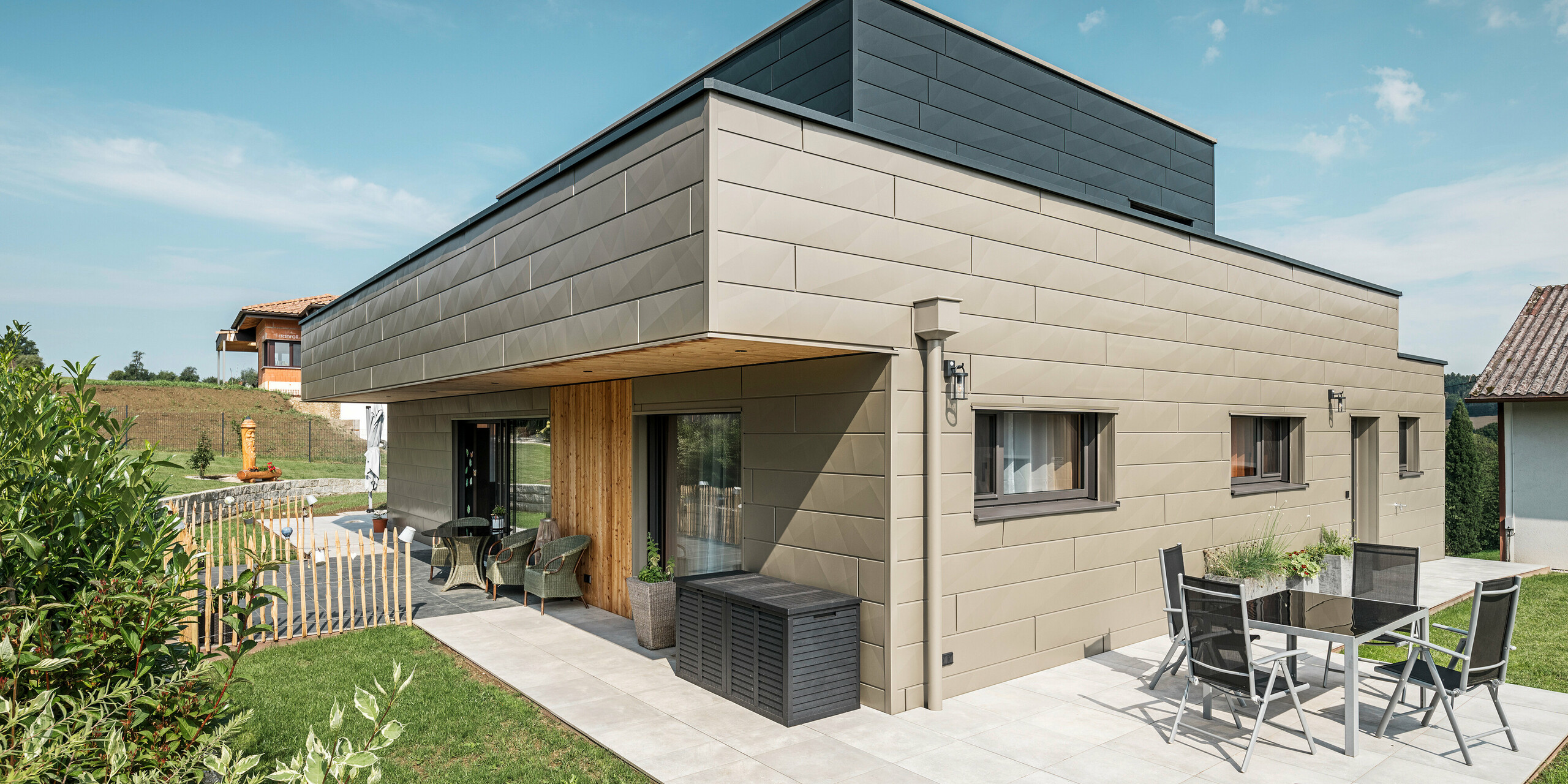 Ein modernes Einfamilienhaus in Wippenham, Österreich, mit einer zweifarbigen PREFA Aluminiumfassade, die aus den extravaganten Siding.X Fassadenpaneelen in Bronze und Anthrazit besteht. Die Fassade zeigt klar erkennbare, elegante Linien und Kantungen. Deutlich sichtbar ist die harmonisch integrierte PREFA Dachentwässerung mit Wasserfangkasten und Ablaufrohr, die sich nahtlos in das architektonische Gesamtkonzept einfügt. Im Vordergrund ist eine gemütliche Terrasse mit Gartenmöbeln zu sehen, die das moderne Design des neugebauten Hauses unterstreicht.