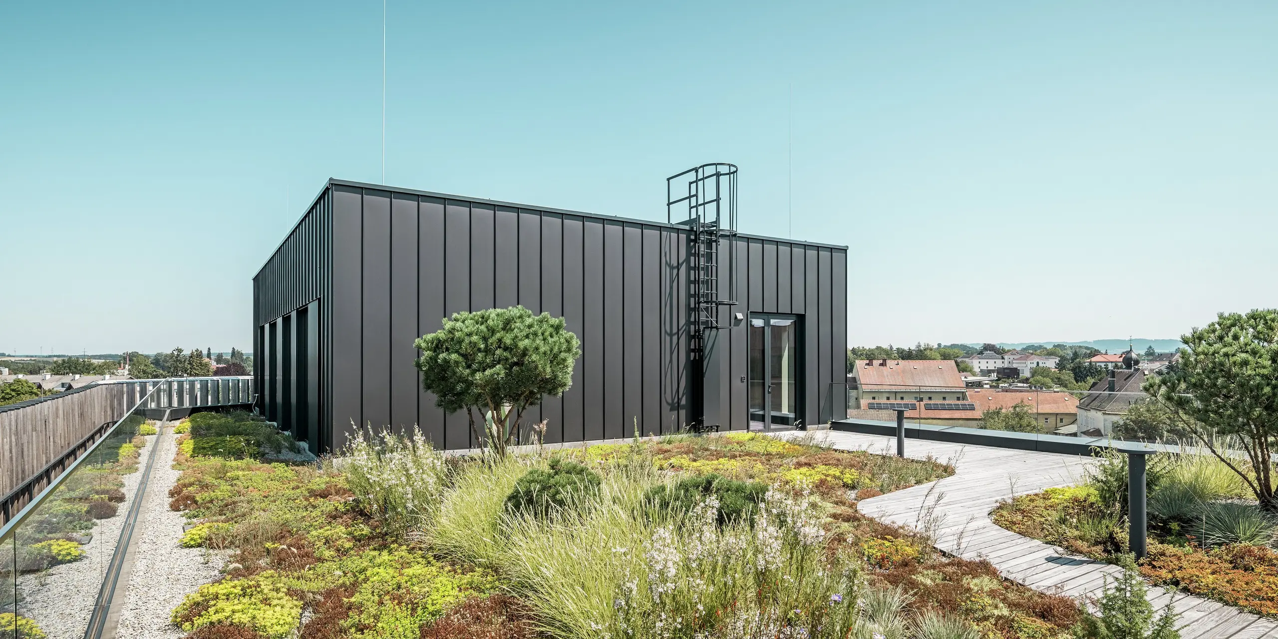 Das begehbare Dach der Team 7 Welt in Ried im Innkreis kombiniert eine schwarzbraune PREFALZ Fassade mit einem begrünten Dach. Die hochwertige Aluminiumverkleidung überzeugt durch Langlebigkeit und Witterungsbeständigkeit, während der Dachgarten ein nachhaltiges und naturnahes Element einbringt. Diese Kombination aus fortschrittlicher Architektur und ökologischem Design schafft einen harmonischen Rückzugsort über den Dächern der Stadt.