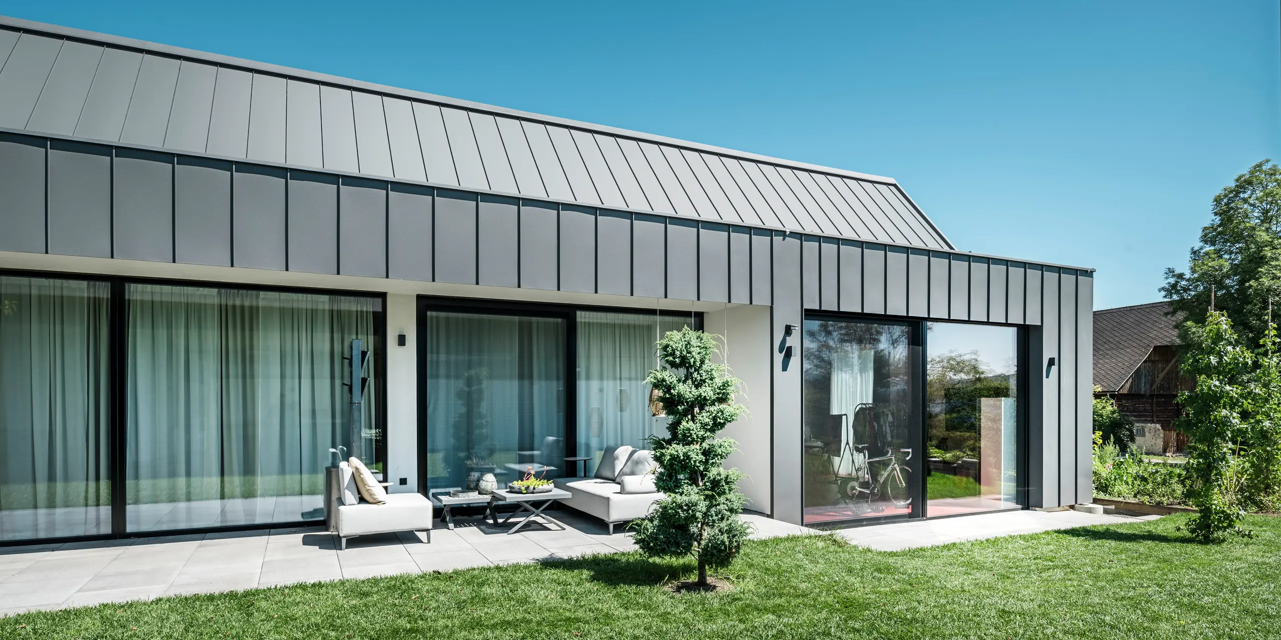 Entspannte Gartenterrasse eines Einfamilienhauses in Pogier, Österreich, mit einer eleganten PREFALZ-Dach- und Fassadenverkleidung in P.10 Dunkelgrau von PREFA. Die großflächigen Glasfronten öffnen den Wohnraum zur Natur und ermöglichen einen fließenden Übergang zwischen Innen- und Außenbereich. Modernes Design trifft auf Funktionalität, wobei die grüne Umgebung und der sorgfältig gepflegte Rasen die ruhige und friedliche Atmosphäre des Ortes unterstreichen.