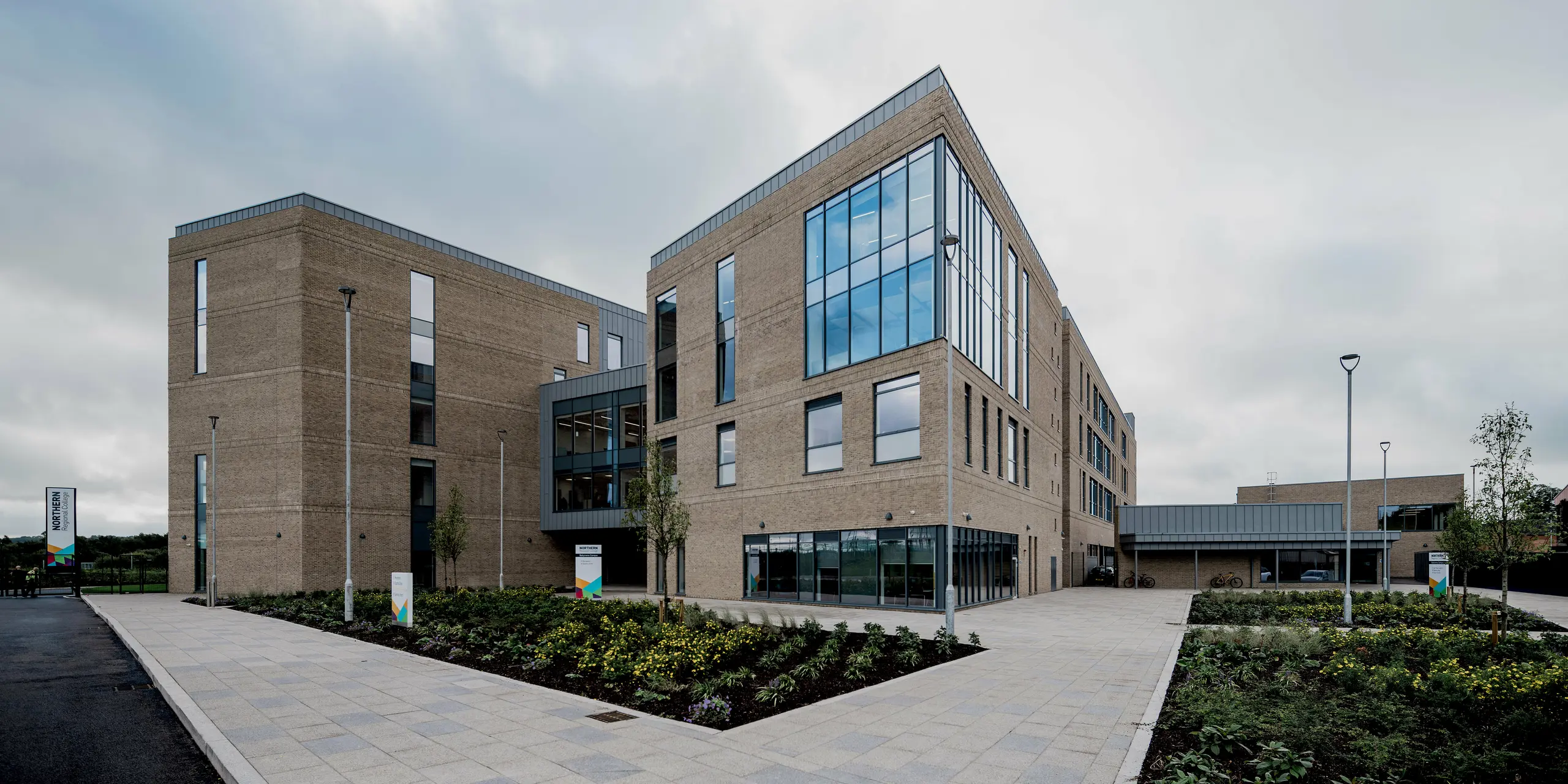 Eine großzügige Gesamtansicht des Northern Regional College in Ballymena zeigt mehrere klar gegliederte Baukörper aus hellem Klinker, verbunden durch Glasflächen und offene Wege. Entlang der Dachkanten und Fassadenabschlüsse setzen PREFALZ Aluminium-Stehfalzbahnen in Patinagrau präzise horizontale Akzente. Die Metallflächen rahmen die Architektur elegant und kontrastieren mit dem Mauerwerk und dem weiten Himmel. PREFA Aluminium überzeugt durch Langlebigkeit, Wartungsarmut und eine zeitlos moderne Gestaltung für Bildungsbauten.
