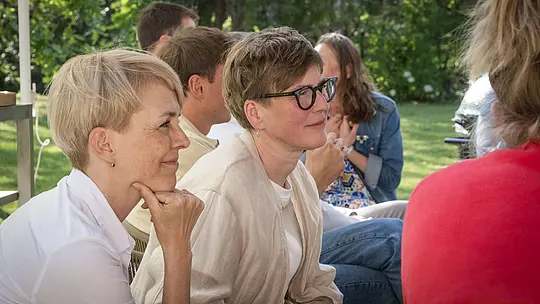 Nahaufnahme von Veronika Roll (PREFA Marketing) und Juliane Croce (Fotografin) bei der PREFARENZEN 2027-Expertendiskussion im Garten einer Winzerresidenz. Die beiden Frauen sitzen konzentriert und aufmerksam und diskutieren lebhaft über moderne Architekturprojekte. Die Szene zeigt natürliche Beleuchtung, sommerliches Grün und eine entspannte Atmosphäre. Im Hintergrund sind PREFA Aluminiumprodukte zu sehen, die langlebige, ästhetisch ansprechende Dächer und Fassaden symbolisieren, bei denen Funktionalität mit Design vereint wird. Das Bild betont den kreativen Austausch und die intensive Auseinandersetzung der Jury mit Designideen sowie die Inspiration, die durch die hochwertigen, innovativen Aluminiumlösungen von PREFA für Architektur und Bauprojekte entsteht.